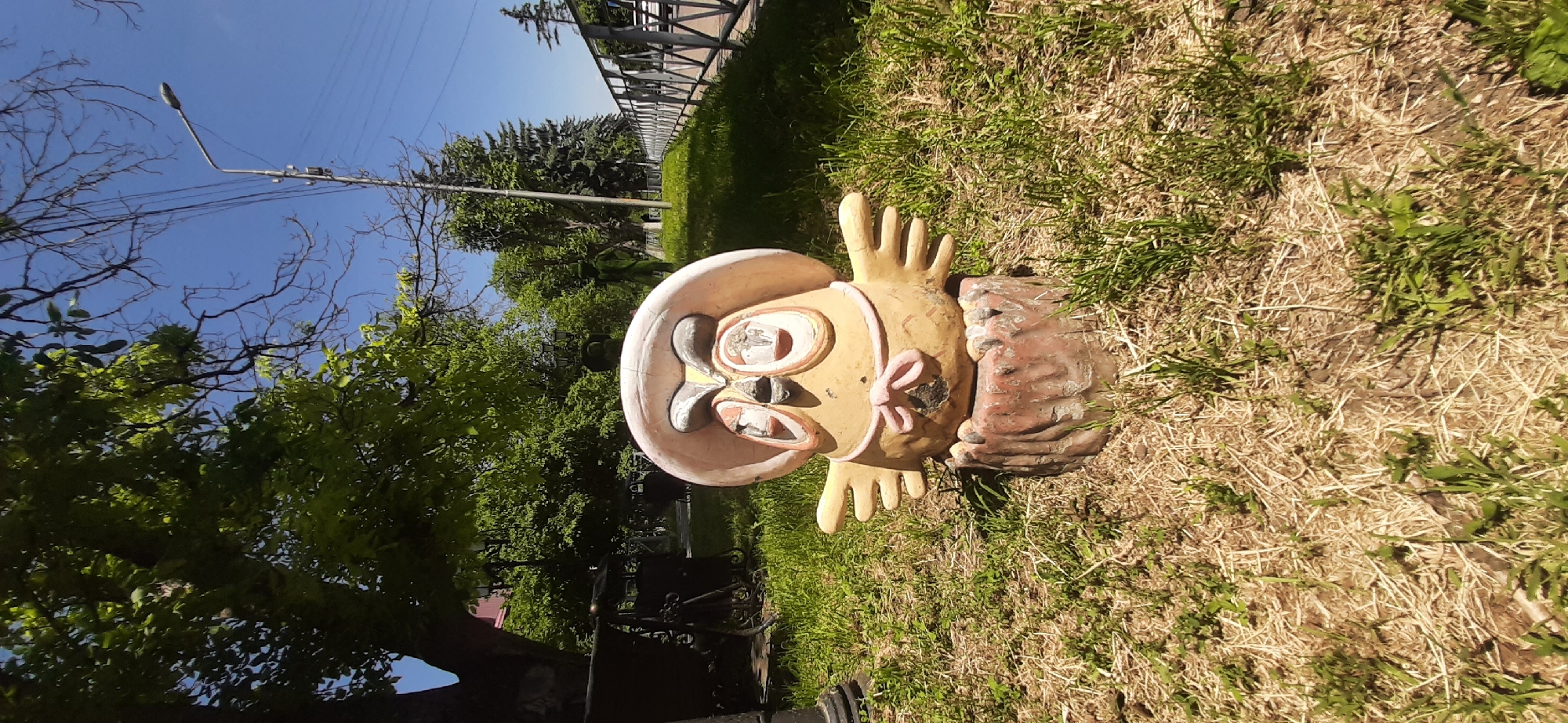 A whimsical, weathered concrete statue of an owl wearing a wide-brimmed hat and a pink bow sits on a reddish-brown, tree-stump-like base in a grassy area. The owl statue is painted in shades of yellow, pink, and white, with large, expressive eyes and outstretched wings. The surrounding environment is a park or public space, with lush green grass, dry straw-like grass, trees, a metal fence, a lamppost, and power lines visible. In the background, a dark metal bench or cart with visible wheels is present. The sky is clear and blue. The scene is located in Yessentukskaya, Russia, indicated by the presence of a lamppost typical of Russian street lighting and the general ambiance of a Russian town's public space.