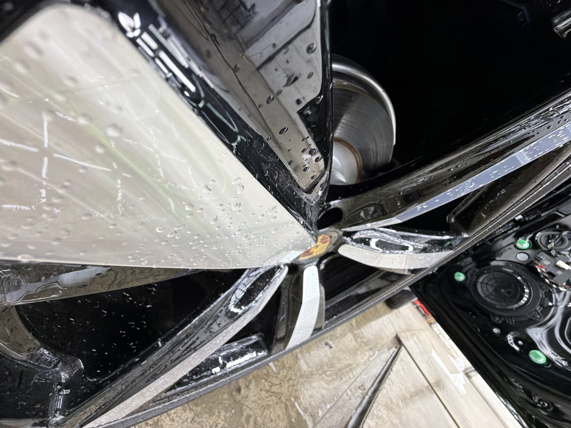 The image depicts the wheel well and driver's side door of a black Porsche, likely a Cayenne, under possible repair or detailing. Visible elements include: the car's multi-spoke alloy wheel with the Porsche crest at the hub, a brake rotor behind the wheel, and the open driver's side door revealing speaker components and wiring. The scene is marked by water droplets suggesting it may have been recently cleaned or is in a humid environment. The setting appears to be an indoor location, possibly a garage or repair shop, due to the neutral-colored floor. There is no evidence in the image to suggest it is in Krasnoyarsk, Russia.