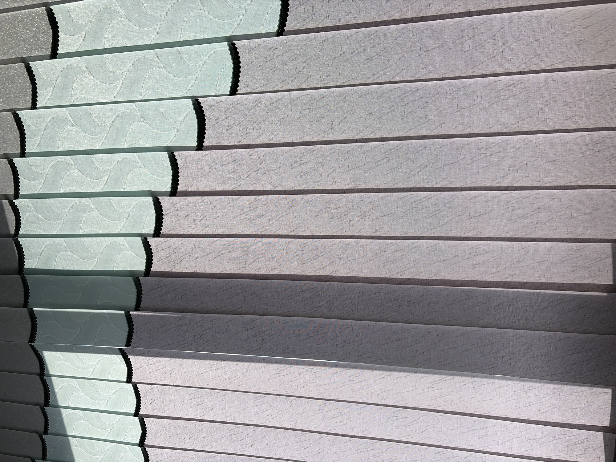 The image depicts a close-up, full-frame view of vertical blinds. The blinds are divided into two horizontal sections. The lower section is a solid, light purple-gray color with a subtle textured pattern. The upper section consists of translucent, light blue fabric with an embossed wavy design. A black, scalloped trim separates the two sections. Sunlight casts strong shadows across the blinds from the left, creating diagonal lines of light and dark. The scene appears to be an interior setting, likely a room with a window covered by these blinds. The location in Podgumer, Bulgaria is not evident from the image itself.