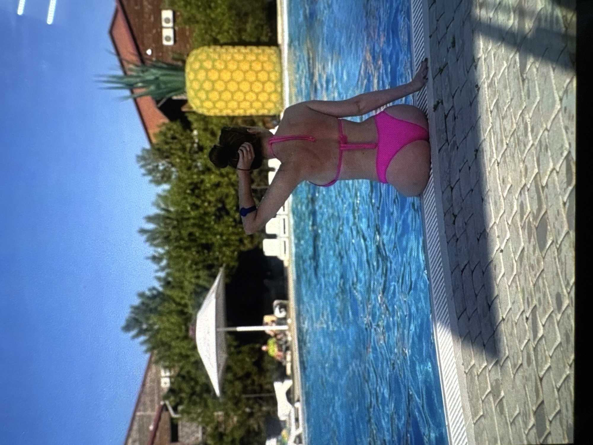 A woman in a pink bikini sits on the edge of a swimming pool, facing away from the camera. She has her right hand on the pool edge and her left hand behind her head, adjusting her hair. In the background, a large inflatable pineapple stands next to a building with a red roof. The swimming pool is filled with bright blue water, and the surrounding area is paved with stone tiles. The sky is clear and blue.
