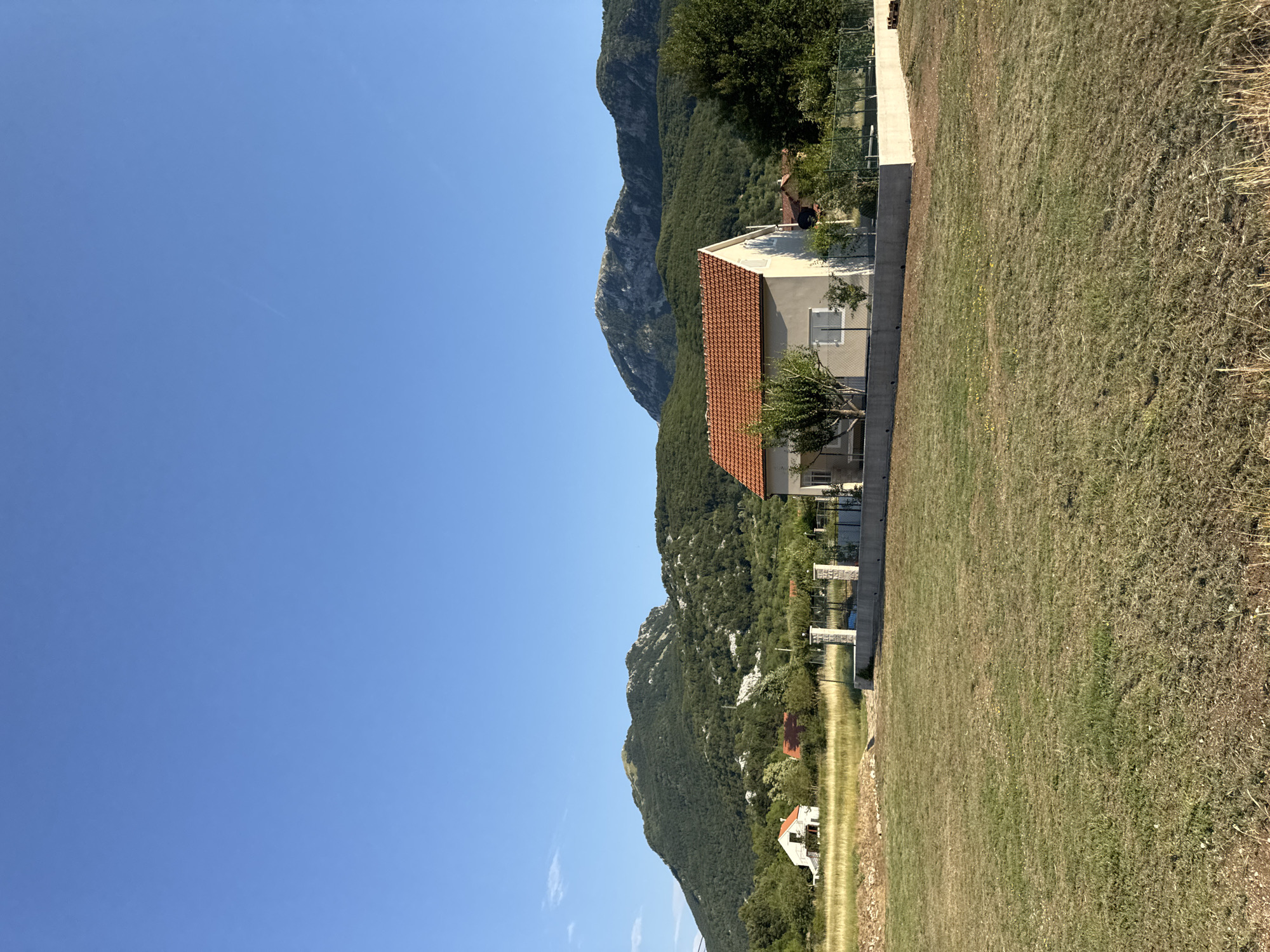 A wide-angle, vertical, outdoor shot shows a residential area in Kopito, Montenegro, with a clear blue sky overhead. In the foreground, a large expanse of dry, patchy grass occupies the majority of the frame. A light-colored house with a red-tiled roof is situated in the mid-ground, right of center, surrounded by a low fence and some greenery. To the left of this house, a smaller white building with a red roof is visible amidst dry grass. In the background, rolling green hills covered in trees and vegetation rise to meet the sky, with rocky peaks visible on the horizon.