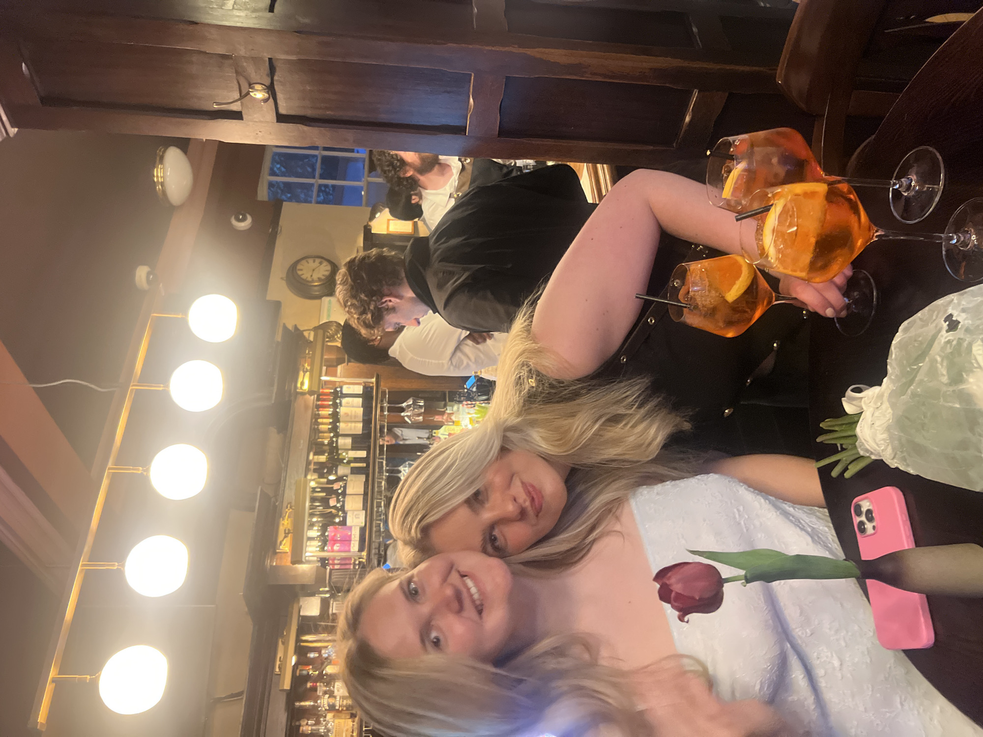 Two young women are posing for a photo in what appears to be a busy pub or restaurant. One woman is wearing a white textured dress, and the other is wearing a black top. On the table in front of them are three glasses of orange cocktails with orange slices, a pink smartphone, and a red tulip. In the background, other patrons are visible, along with a well-stocked bar featuring numerous bottles. The lighting is warm, suggesting the use of incandescent bulbs. A clock is on the wall above the bar. The architecture suggests a traditional British pub setting in the city of London, United Kingdom.