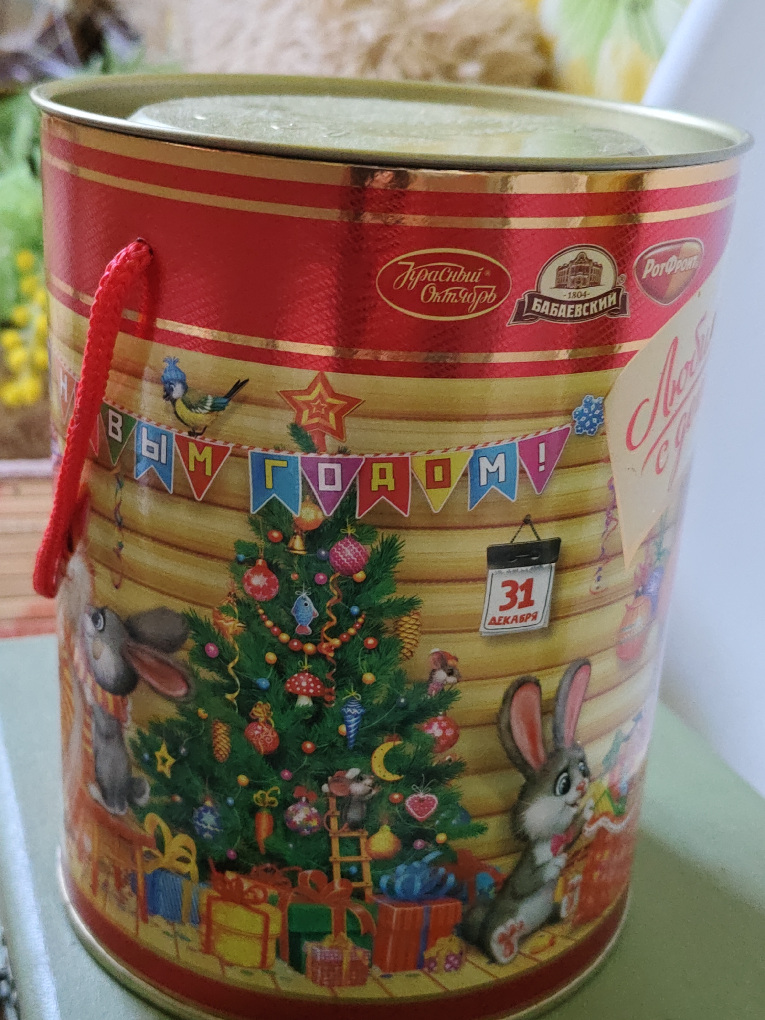 A cylindrical tin can, decorated with a Christmas theme, is the subject of the image. The can features a bright red band with gold stripes at the top, below which is a yellow background with a wood-like texture. Adorning the yellow surface are illustrations of a Christmas tree, presents, rabbits, a bird wearing a blue hat, and a banner with the Cyrillic text "С НОВЫМ ГОДОМ!" (Happy New Year!). A small calendar displays "31 ДЕКАБРЯ" (December 31). A red rope handle is attached to the top left of the can. There are also logos on the upper part of the can, including "Красный Октябрь" (Red October) and "Бабаевский" (Babaevsky). The can is placed on a light green surface. The background is blurred, showing hints of greenery and yellow flowers. There is no discernible location in the city of Rostov-on-Don, Russia, or any people present in the image.