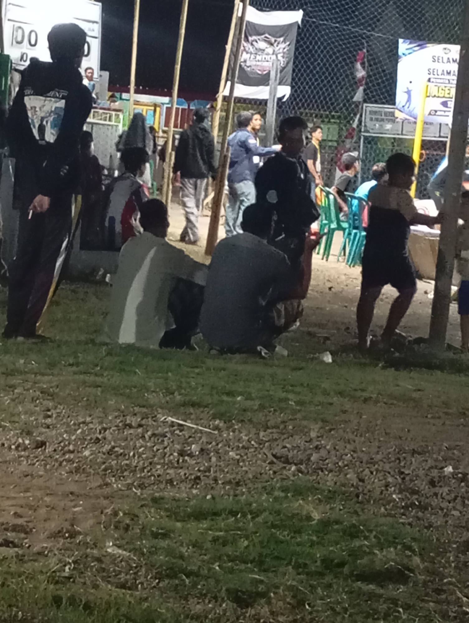 The image depicts an outdoor scene at night, likely a community event or gathering, possibly in Pacitan, Indonesia, given the Indonesian text visible on banners. In the foreground, a grassy area transitions to a dirt/gravel path. Several individuals are present, some standing and some seated. One person in the foreground, partially obscured, is wearing dark clothing and holding a cigarette. To their right, another person is squatting and looking towards the right. In the midground, more people are gathered, with some standing near chairs. A banner with the text "SELAMAT..." is visible in the background. Another banner with a logo and the word "MENDO" is also present. The scene appears to be dimly lit, suggesting it is nighttime. The overall impression is of a casual social gathering in a semi-public space.