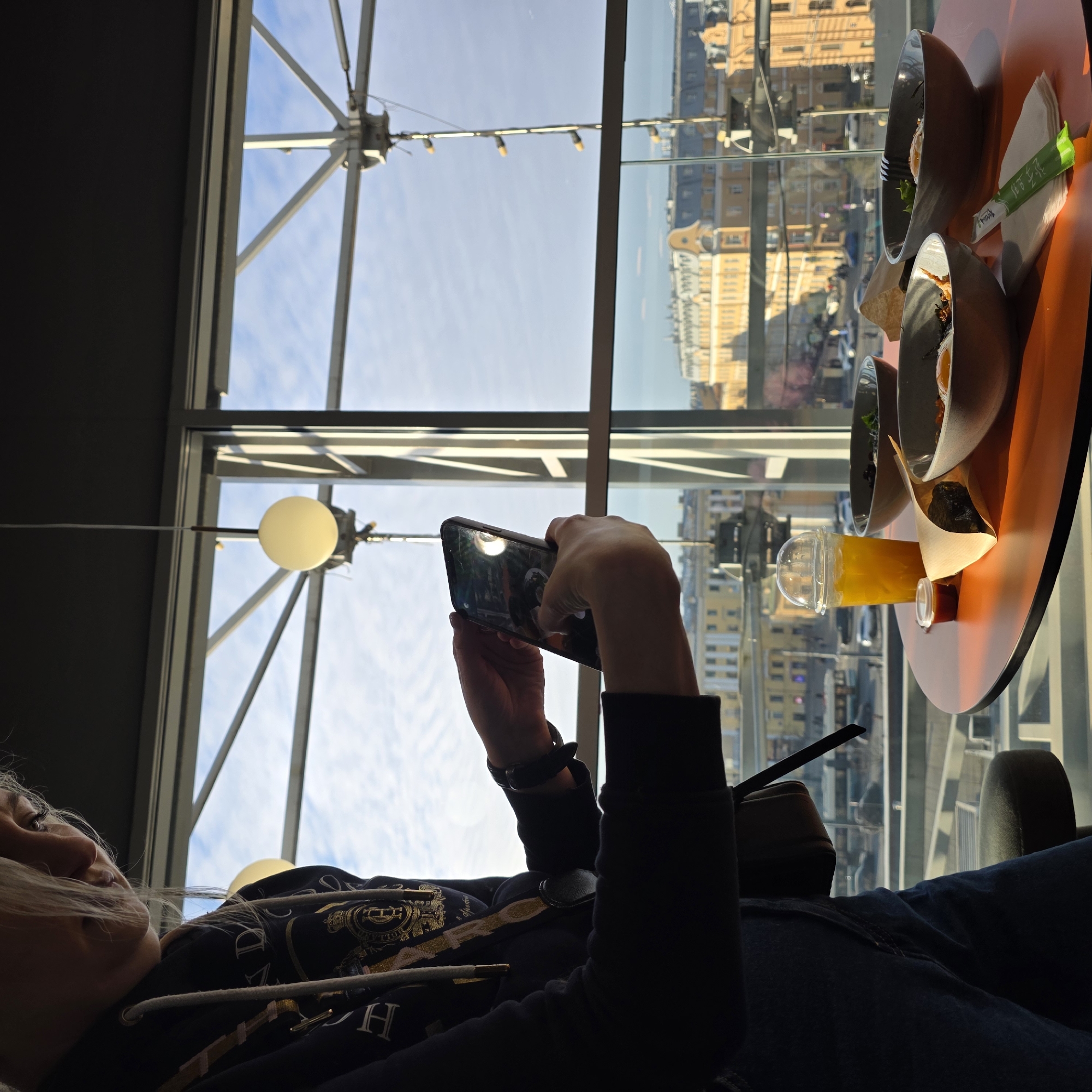 A woman is sitting in a chair by a window in Unknown, Unknown. She is wearing a black hoodie and jeans, and she has a black wristwatch. The woman is holding a smartphone in her hands and appears to be taking a picture. On the table next to her are several bowls of food, a drink, and chopsticks. The window offers a view of the city skyline. The building has a visible support structure, and there are hanging light fixtures.