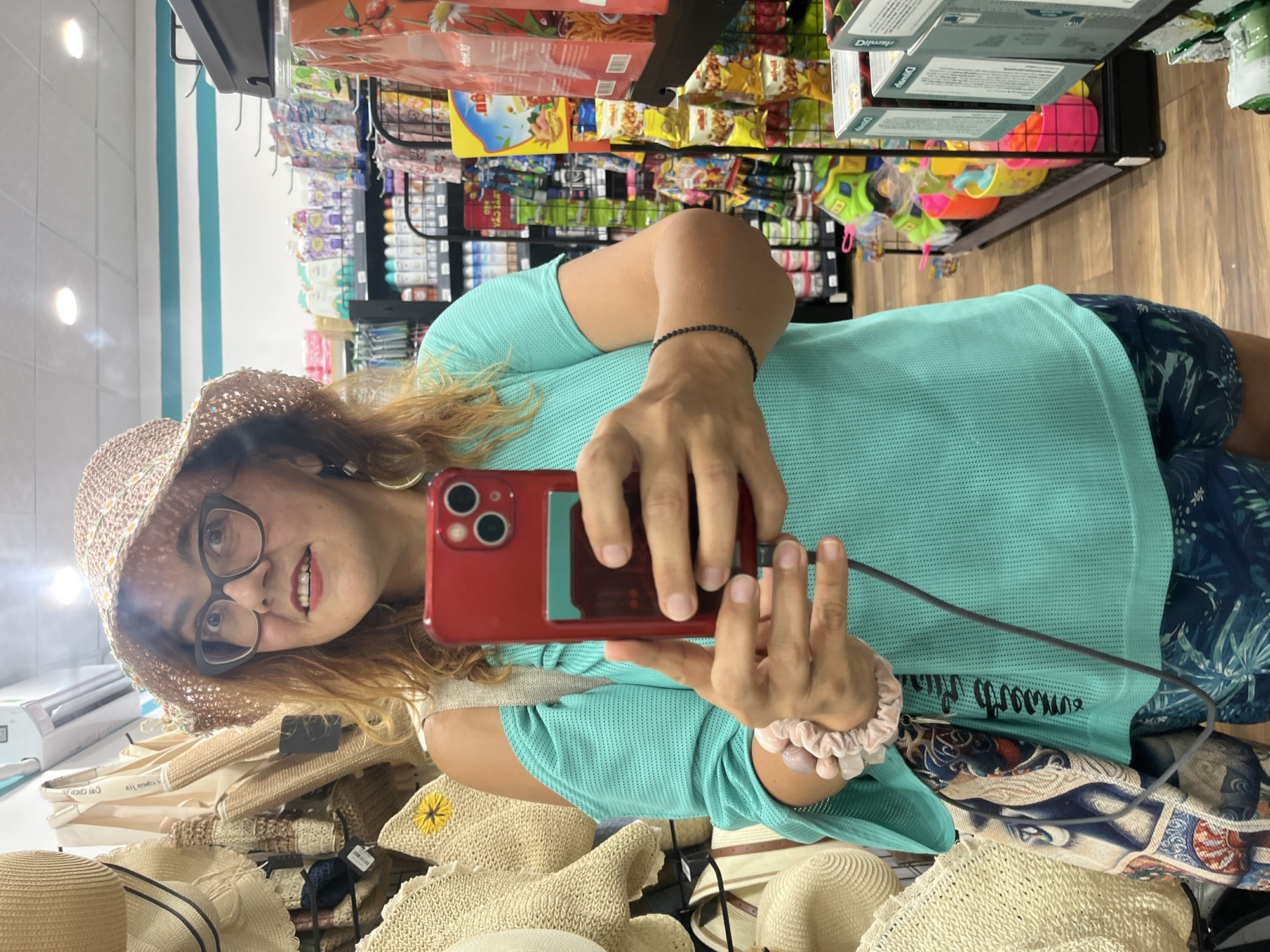 A woman is taking a selfie in a store. She is wearing a straw hat with daisy trim, glasses, and a turquoise off-the-shoulder top. She is holding a red smartphone in her right hand, which is angled to capture her reflection. In her left hand, she holds the phone's charging cable. Her left wrist is adorned with a pink scrunchie and a bracelet made of light pink beads. A patterned scarf is draped over her left shoulder, and she is wearing dark blue patterned shorts. The background shows shelves stocked with various items, including hats, bags, toiletries, and snacks. The scene appears to be in a retail environment within Nha Trang Ward, Vietnam.
