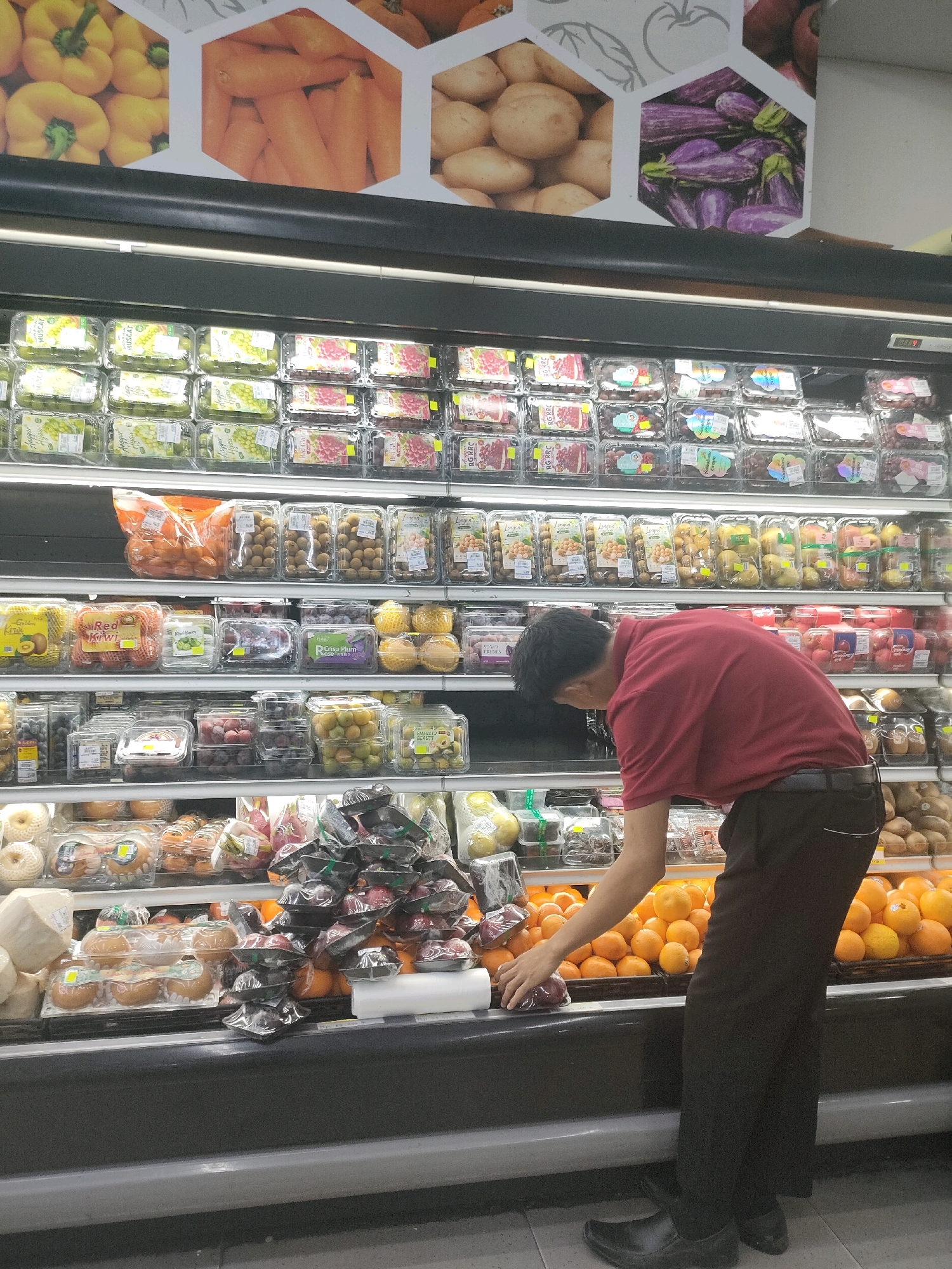 A man in a red shirt and dark pants is seen from the side in a grocery store produce aisle. He is reaching into a display of oranges. The shelves behind him are stocked with various fruits and vegetables, including grapes, kiwis, apples, and what appear to be dragon fruits. The store is likely a supermarket chain common in Batam, Indonesia, with brightly lit displays and organized packaging of produce. The image appears to be taken during daylight hours.
