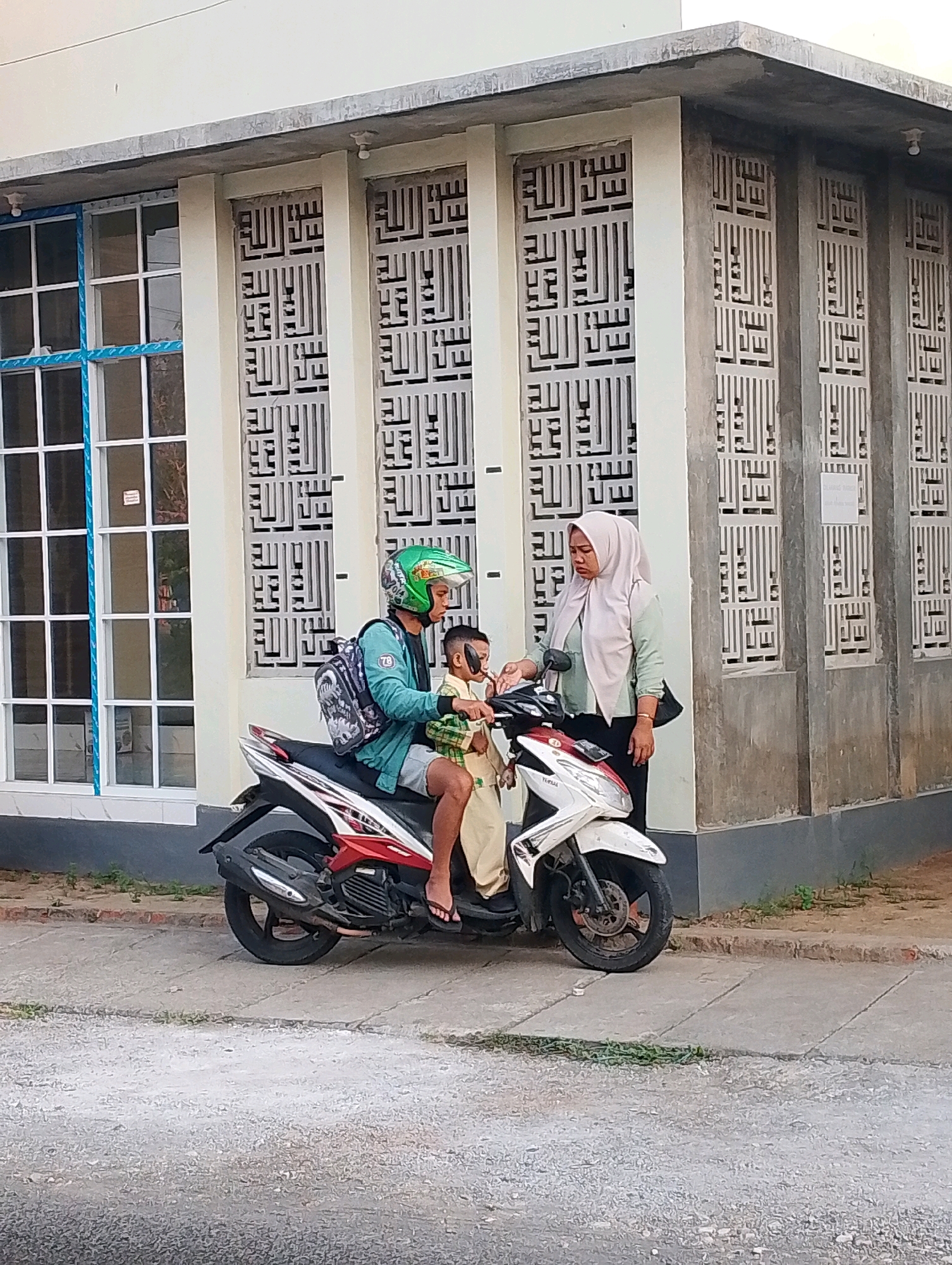 An adult male wearing a green helmet and a blue jacket sits on a scooter with a young boy in front of him, also on the scooter. A woman wearing a light-colored hijab and a green top stands next to the scooter, appearing to interact with the boy. The scooter is parked on a paved surface next to a building with decorative panels featuring Islamic calligraphy. A large window with a grid pattern is visible on the left side of the building. The background suggests an outdoor, urban setting. The image was captured in Probolinggo, Indonesia.