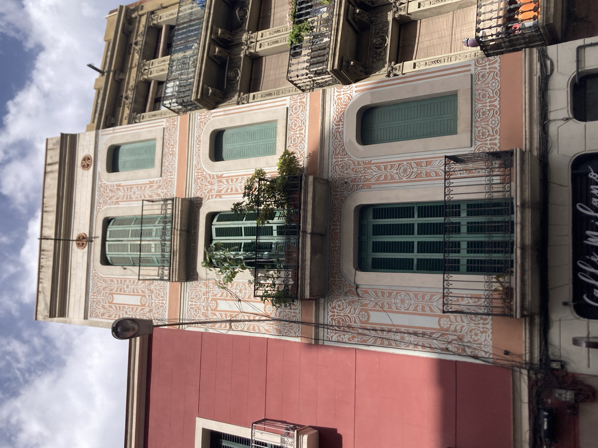 This is a low-angle shot of a colorful street in Barcelona, Spain, under a cloudy sky. On the left, a building with a vibrant pink facade has a simple white-trimmed window and a small wrought-iron balcony. To its right, a narrower building features a light peach-colored facade adorned with intricate white relief patterns of floral motifs and vertical stripes. This building has three floors visible, each with a green shuttered window and a wrought-iron balcony. The balconies on the second and third floors hold potted plants. To the far right, another building with a yellow-tan facade is visible, also featuring multiple balconies with ornate wrought-iron railings. A street lamp stands on the left, its head angled downwards. Wires are strung between the buildings. At the bottom of the frame, the sign for "Caffè Milano" is partially visible on a storefront. The overall impression is one of architectural diversity and lively urban life.