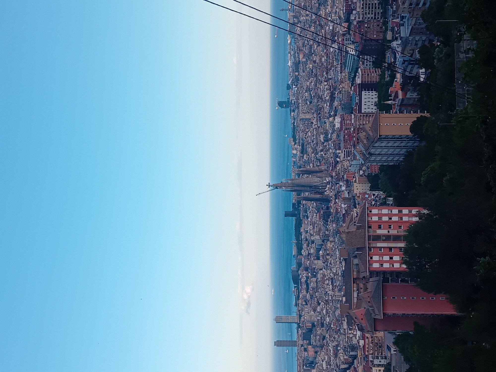 The image depicts an elevated view of Barcelona, Spain, with the Mediterranean Sea on the horizon. The skyline is dominated by the Sagrada Familia, identifiable by its distinctive spires. A dense urban sprawl fills the space between the viewer and the sea, consisting of closely packed buildings with varying heights and architectural styles. Cables, presumably from a cable car or similar structure, are visible in the upper portion of the frame. Vegetation, likely trees, appears in the foreground, partially obscuring the view on the right side of the image. The sky is a gradient of light blue, indicating daytime conditions.