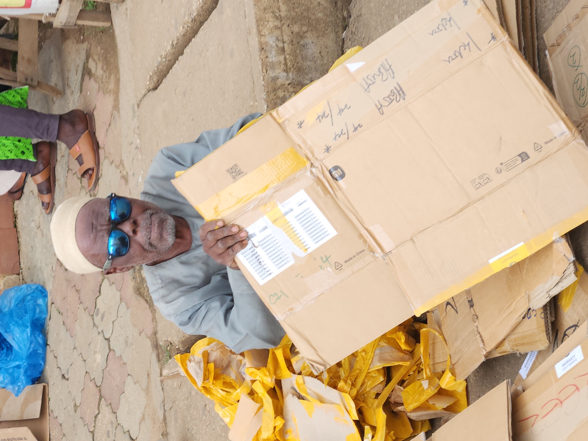 The image shows an elderly African man wearing a light-colored kufi and blue mirrored sunglasses. He is seated outdoors on a paved surface, surrounded by piles of yellow packaging material and cardboard boxes. The man is holding a large, opened cardboard box in front of him, which has handwriting and shipping labels on it. His right hand is resting on the box. He is wearing a grey long-sleeved garment. In the background, parts of other people's legs and footwear are visible, indicating a public space. The ground is a mix of paved tiles and dirt. The overall impression is one of commerce or distribution in an outdoor market or roadside setting.