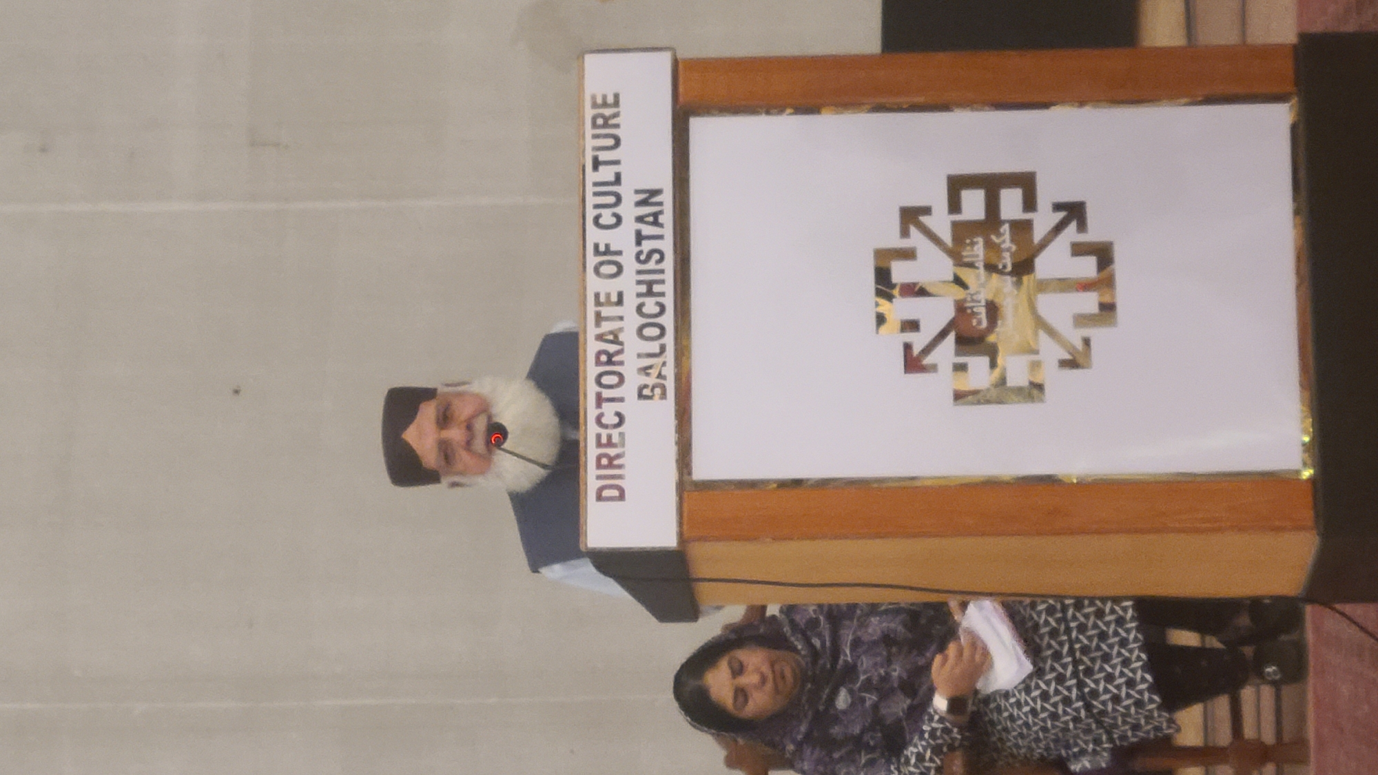 The image shows a man with a white beard and a black cap speaking at a podium. The podium has the words "DIRECTORATE OF CULTURE BALOCHISTAN" printed on its front panel. It also features a symbol in gold color, appearing to be an emblem or logo. A woman wearing a patterned headscarf and blouse is seen below the podium, appearing to be holding a piece of paper. The event seems to be taking place indoors, possibly in a location within the city of Quetta City Tehsil, Pakistan.