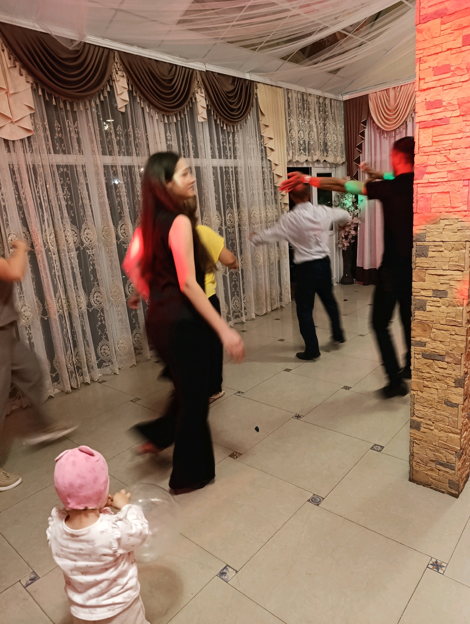 In Korzhevo, Moldova, within a room decorated with brown and beige drapes and sheer white curtains, several people are observed dancing. A child in a pink hat and white pajamas is in the foreground, holding a transparent balloon. To the left, a person in gray pants is moving. In the center, a woman in a black sleeveless top and black pants is dancing. Behind her, a person in a yellow shirt is also dancing. Further back, two men are engaged in dancing, one in a white shirt and dark pants, and another in a black shirt and dark pants. The room has a tiled floor and is illuminated by red lighting from the right side, emanating from a stone-clad pillar. The overall atmosphere suggests a celebratory event or party.
