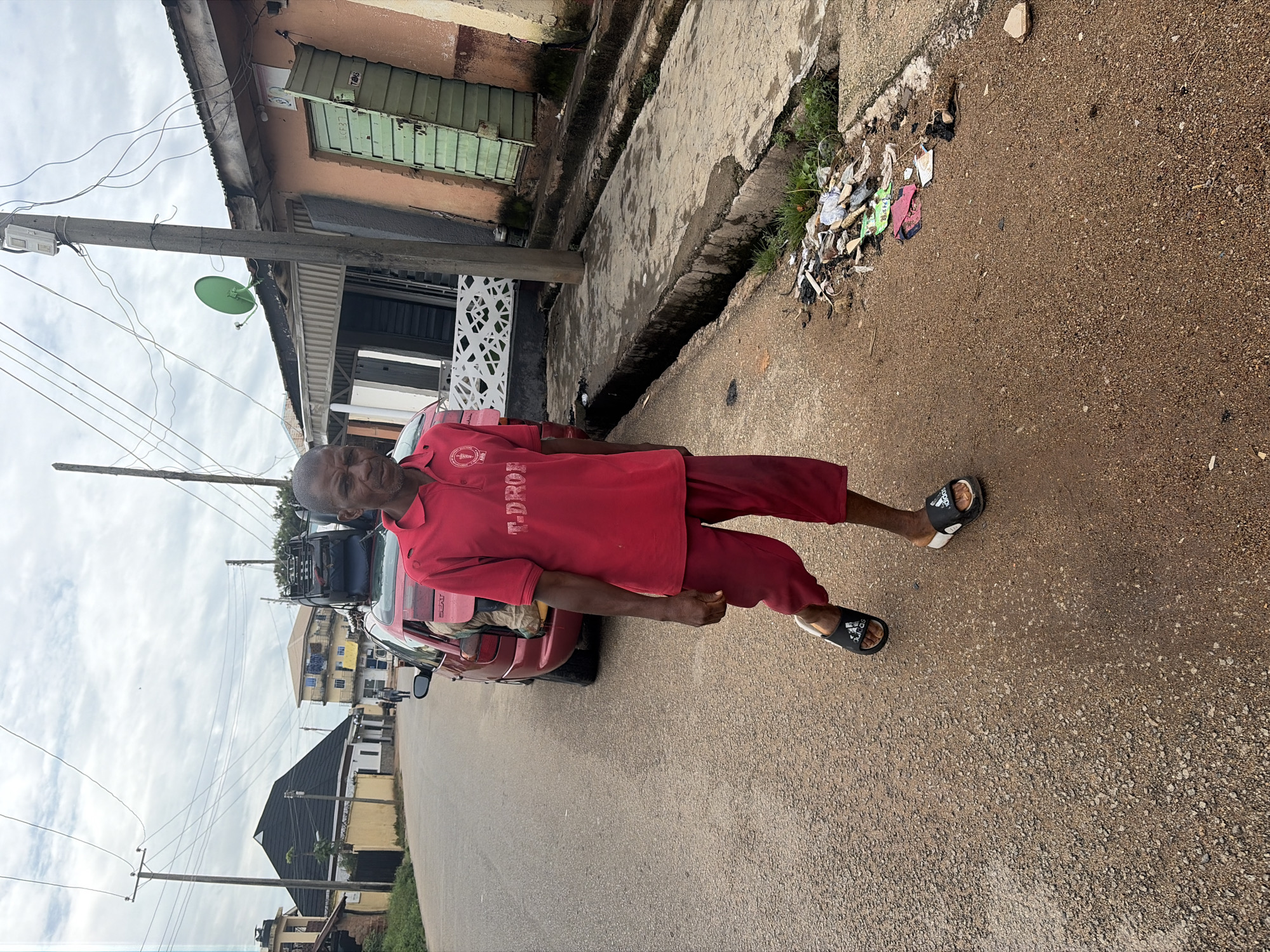 A man wearing a red polo shirt with "T-DROP" printed on it and red shorts walks on a paved road in Jos, Nigeria. He is wearing black Adidas flip-flops. A red car with luggage strapped to its roof is parked on the side of the road next to him. The car's back window is slightly open, revealing a person inside. Buildings line the street, and electrical wires crisscross overhead. A green satellite dish is mounted on the side of one building. The ground on the side of the road is littered with trash.