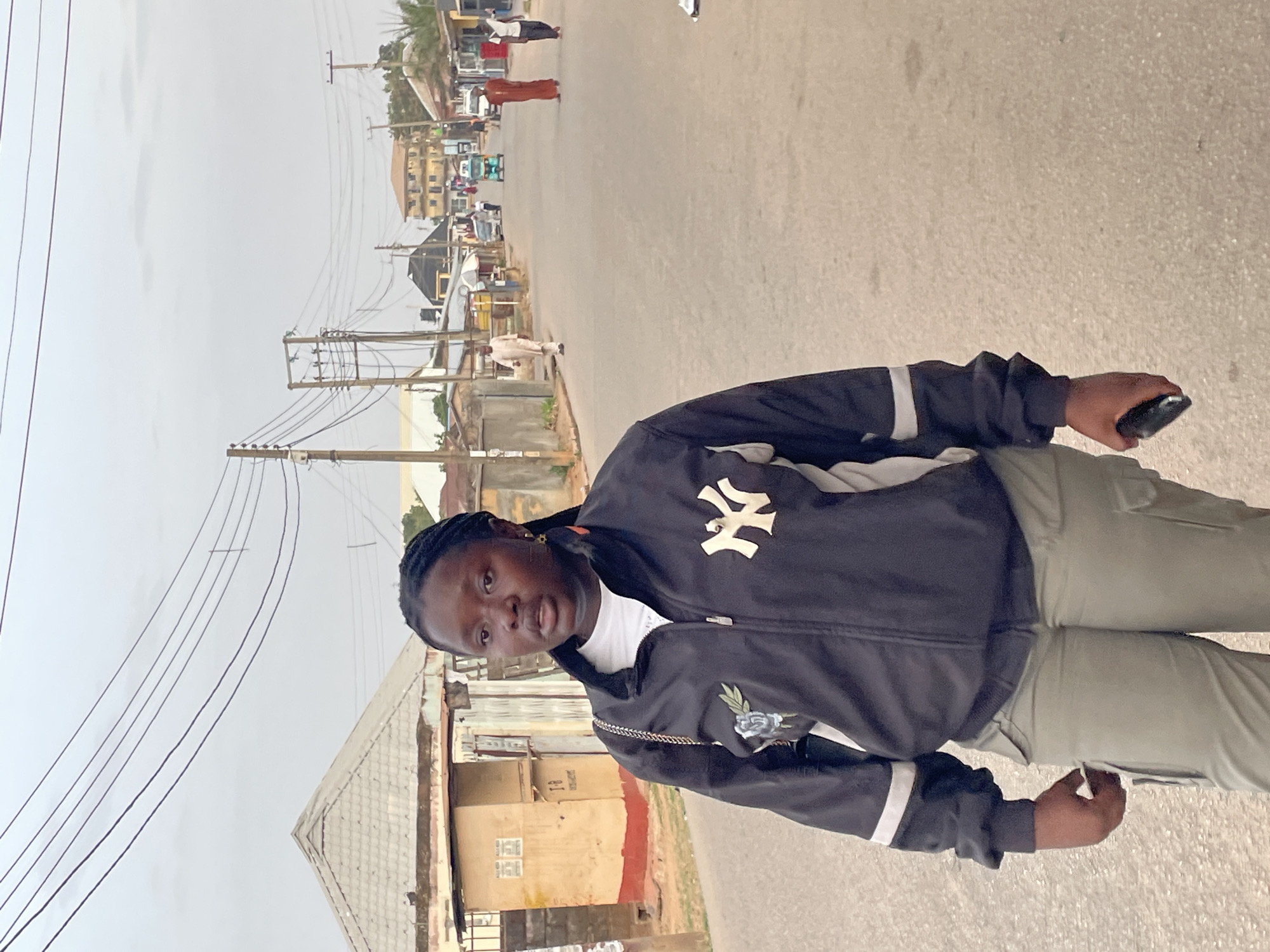 A person stands on a paved road in an urban environment. The person has dark skin, braids, and wears a black jacket with a white NY logo and a floral emblem, a white shirt, and light green pants. The person holds a dark object in their right hand. The background includes buildings, power lines, and other people on the road. The sky is overcast.