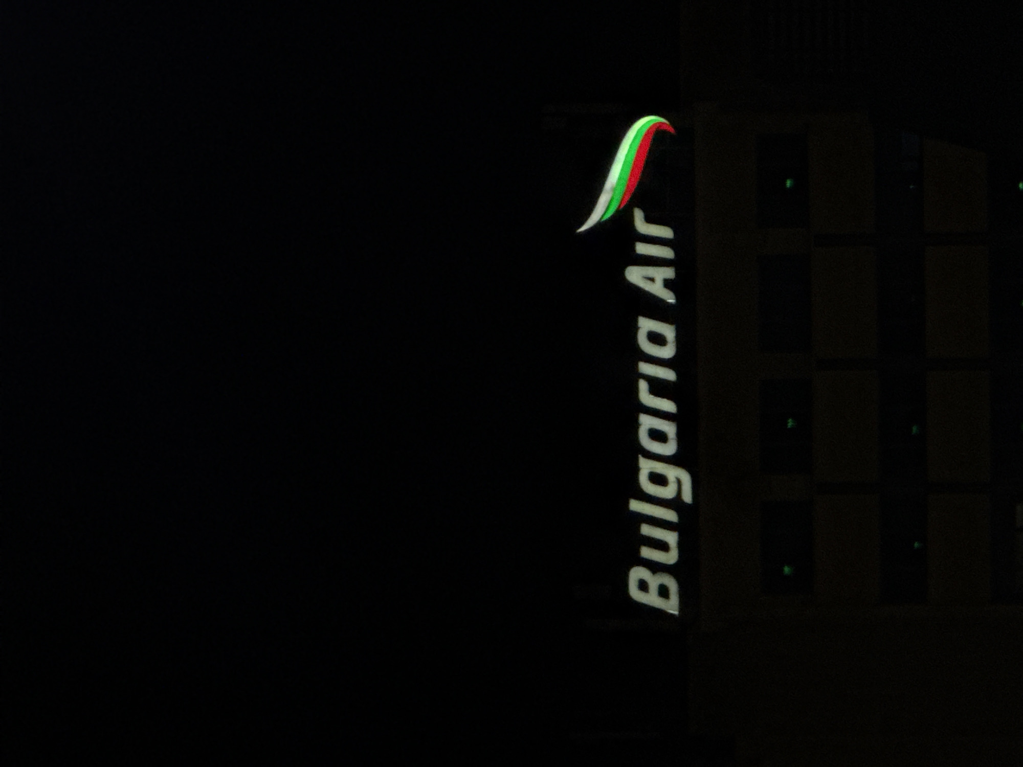 The image displays an illuminated vertical sign for "Bulgaria Air" set against a dark night sky. The sign, positioned on the right side of the frame, features the airline's name written in white capital letters, oriented vertically. Above the text is the Bulgaria Air logo, a stylized wing or ribbon design composed of green, white, and red stripes, reflecting the colors of the Bulgarian flag.
Behind and to the right of the sign, a dark building facade is dimly visible, showing a pattern of square-shaped window or panel features. The vast majority of the image, particularly the left and central portions, is entirely black, accentuating the night-time setting. No people or specific activities are present, with the focus solely on the brightly lit sign. The time of day is clearly night, and the weather conditions cannot be determined beyond the clear visibility of the sign's illumination. The visible text and logo strongly indicate a location related to the airline in Sofia, Bulgaria, likely an airport terminal or an office building.