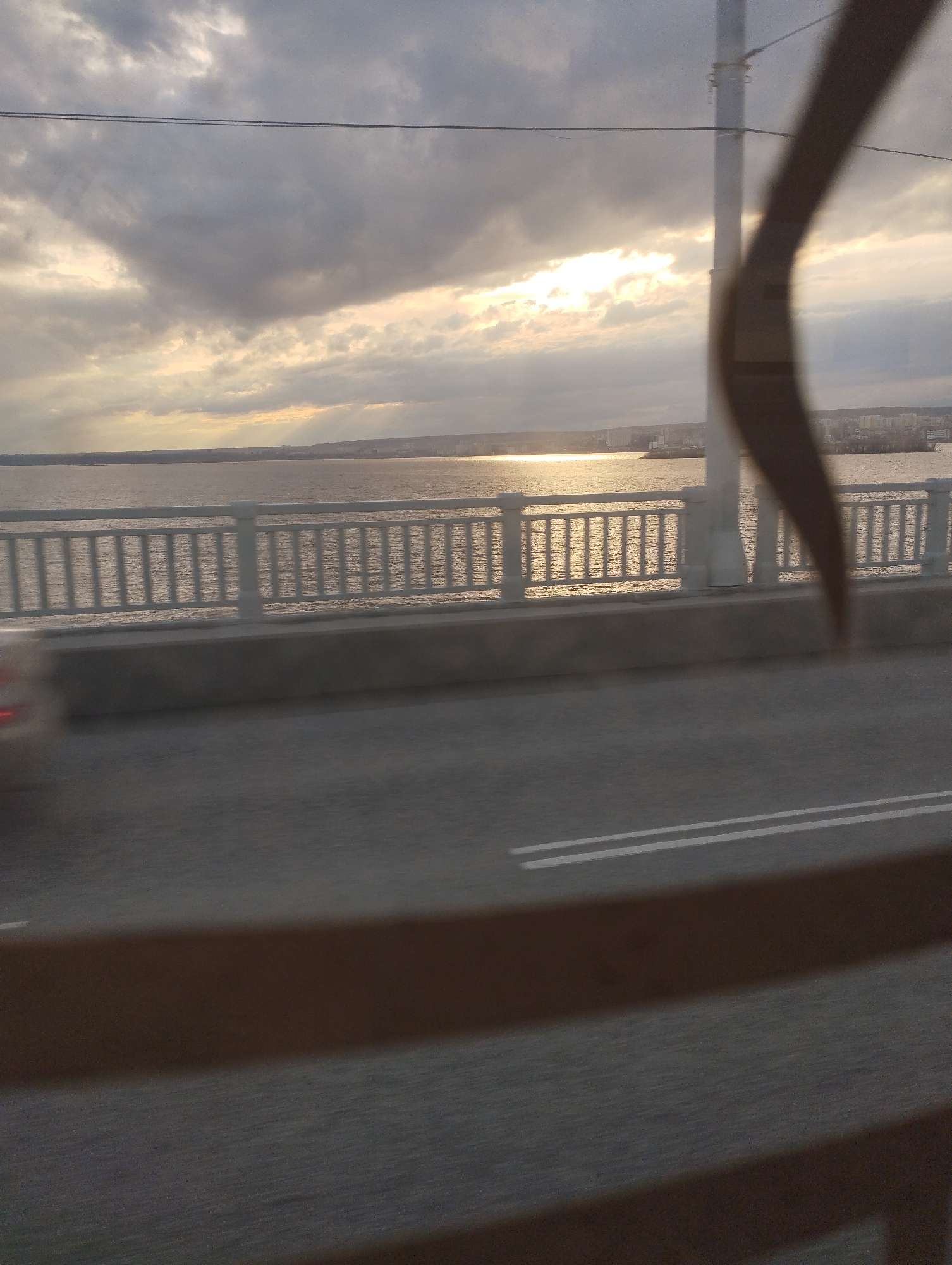 The image shows a view from a vehicle, possibly a bus, traveling on a bridge. The bridge has a gray concrete barrier and a white metal railing. Beyond the railing is a body of water with sunlight reflecting on its surface. On the opposite shore, there is a cityscape. The sky is overcast with clouds, and the sun is obscured. The road surface in the foreground is gray with white lane markings. A blurred object is partially obstructing the view on the right side.