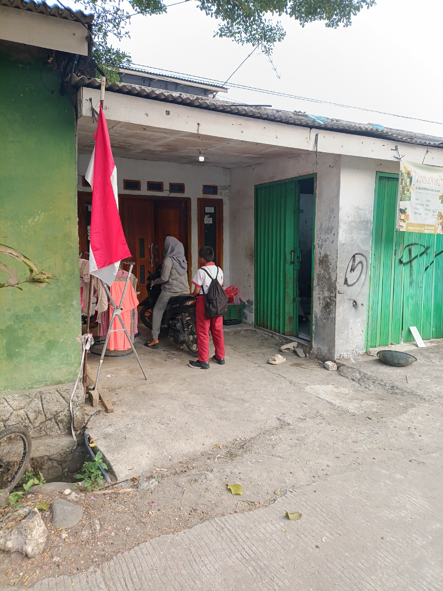 In the city of Cariu, Indonesia, a flag with red and white stripes, possibly the Indonesian flag, is displayed on a metal stand in front of a building. The building appears to be a small shop or residence with green metal roll-up doors. A woman wearing a hijab and a grey top sits on a motorcycle. Next to her, a young boy with a backpack stands, facing away from the camera. Clothing items are displayed on a rack to the left of the flag. The ground is paved with concrete and appears weathered. In the background, trees can be seen.