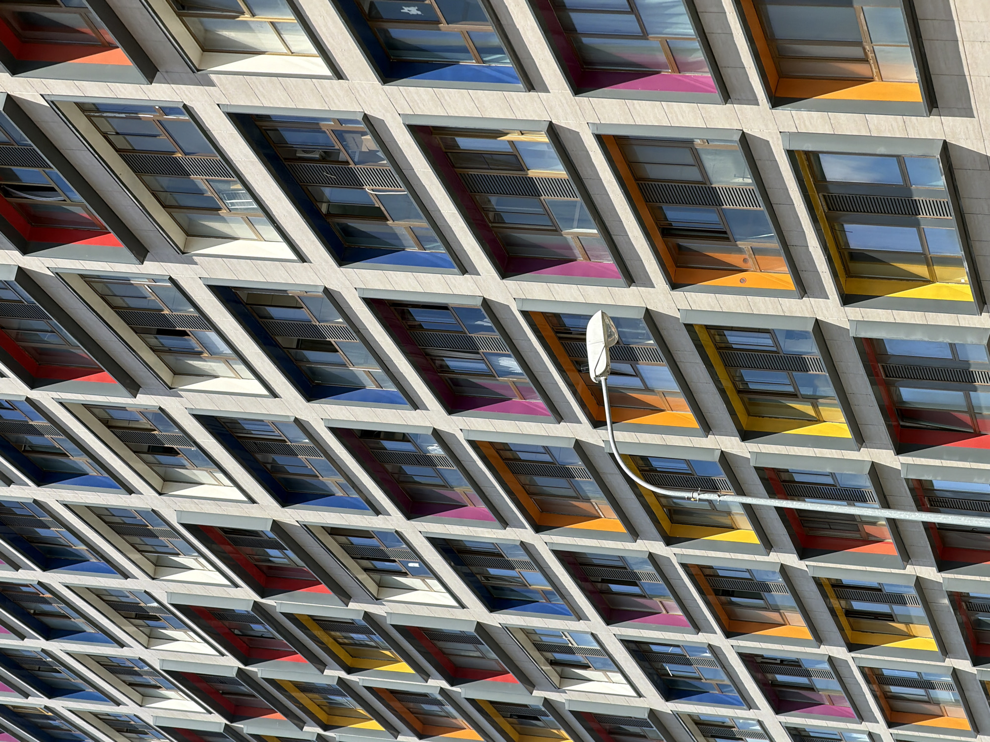 This is a medium-angle, high-contrast photograph of a building's exterior, showcasing a repeating pattern of angular windows set into a light-colored, textured facade. The windows are arranged in diagonal rows, giving the building a geometric and almost honeycomb-like appearance. Each window unit appears to be a deep recess containing multiple panes of glass and dark slatted ventilation grates.
The back interior of some of these recesses are painted in bright, saturated colors, including red, yellow, purple, and orange, creating a striking contrast with the cool tones of the glass and the neutral building material. The colored sections are sharply defined, suggesting a deliberate architectural design choice.
The light suggests it is daytime, with strong shadows emphasizing the depth and angles of the building's structure. The sky is not visible, so weather conditions cannot be determined, but the bright sunlight indicates a clear day. There is a curved pipe or conduit running horizontally across the middle of the image, attaching to the building with a bracket. This appears to be a functional element, contrasting with the aesthetic design of the windows.
No people are visible in the image. The focus is entirely on the architectural elements of the building. There is no visible text in the image. The overall impression is one of modern, bold architectural design with a playful use of color.