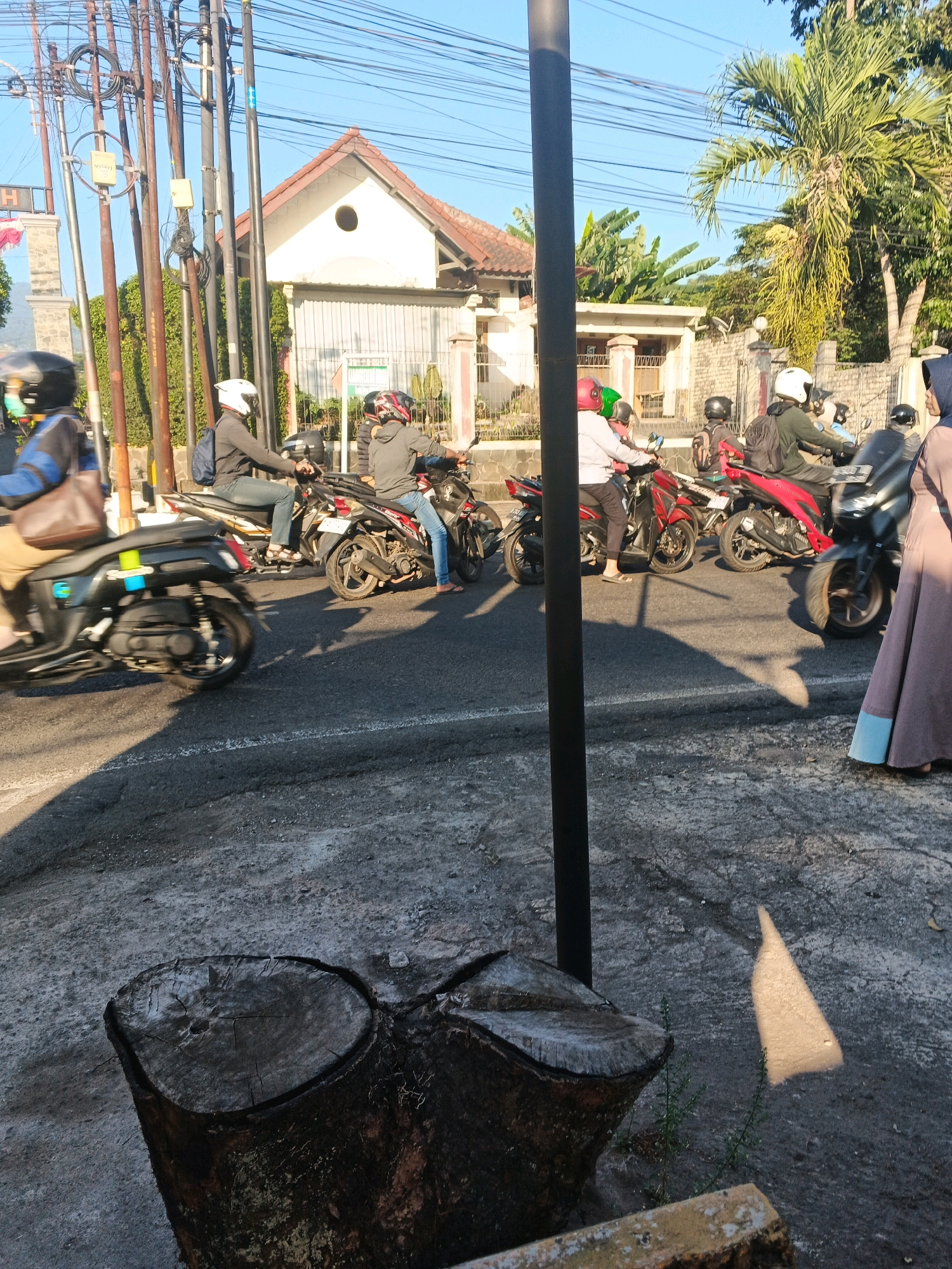 The image depicts a street scene in Semarang, Indonesia, with several motorcycles and riders navigating a road. The foreground features a section of rough pavement and a tree stump, suggesting an urban outdoor environment. In the middle ground, a line of motorcycles is visible on the asphalt road. One motorcycle in the foreground is black and appears to be in motion. Behind it, several other motorcycles are stationary or moving slowly, with riders wearing helmets. The riders are diverse in their attire, some wearing casual clothing while others are in more protective gear. The background reveals a building with a distinctive terracotta-tiled roof and a white facade, characteristic of Indonesian architecture. There are also power lines and vegetation, including palm trees, which are common in tropical urban settings. The lighting suggests it is daytime, possibly late afternoon, given the shadows cast on the ground. The overall atmosphere is that of a typical day in a moderately busy Indonesian city.