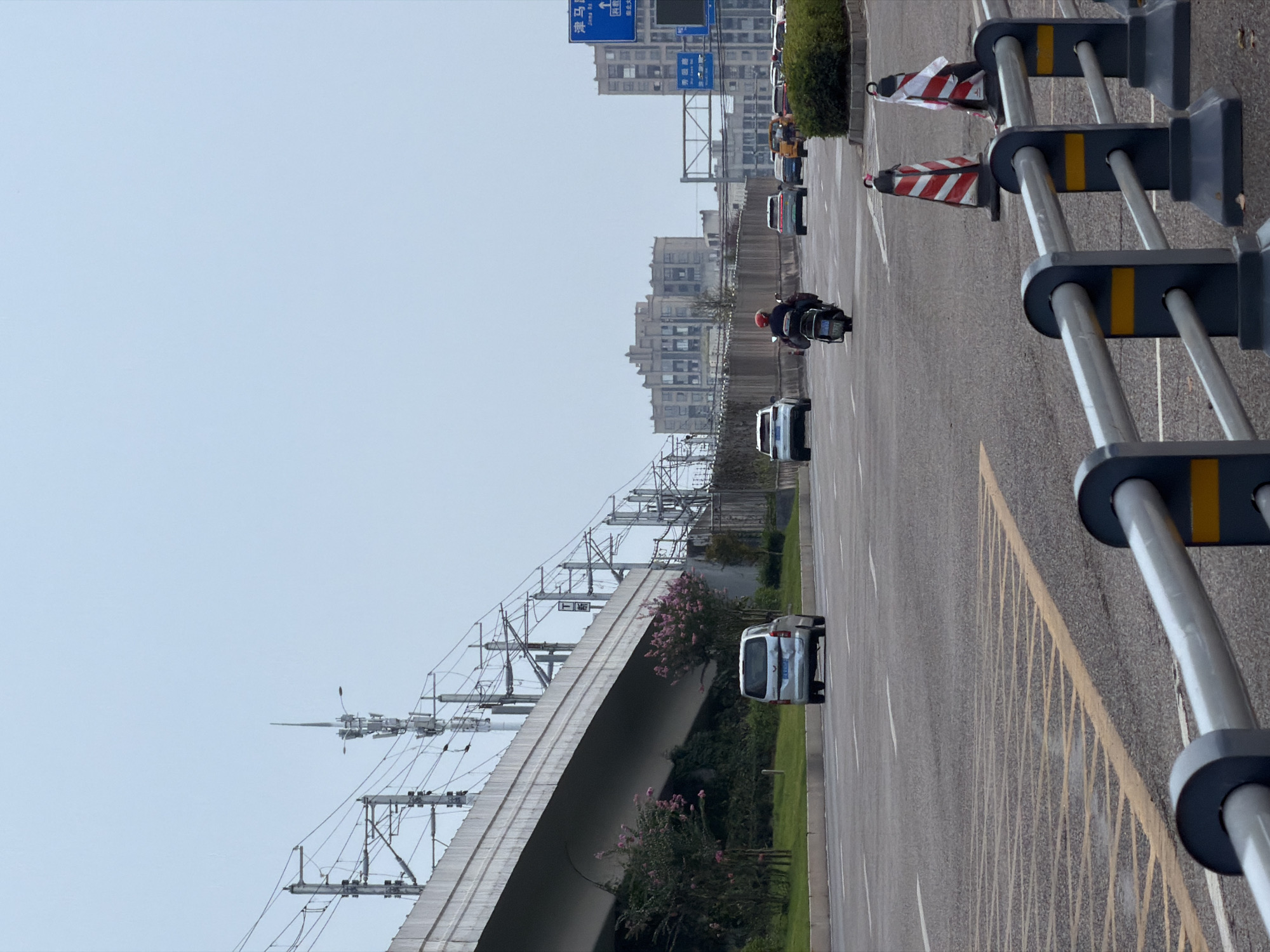 The image captures a wide, multi-lane road in Jiangjin, China, under a hazy, overcast sky. A raised railway or highway overpass dominates the left side of the frame, supported by concrete structures and laden with overhead electrical lines for trains. Below the overpass, a line of trees, some with pink blossoms, lines the grassy median and embankment.
The road itself is asphalt, marked with white lane dividers and yellow hatching in the foreground. Traffic is moderate, with several vehicles visible. A silver minivan is seen in the left lane of the overpass, moving away from the viewer. Further down the road, a person on a black motorcycle, wearing a red helmet and a backpack, is in the center lane, also moving away. Several other cars, including white SUVs and a yellow taxi, are dispersed in the traffic. In the right lane, a row of traffic cones, marked with red and white stripes, signals a potential lane closure or hazard.
In the background, multi-story, beige-colored buildings with repetitive window patterns suggest an urban or suburban setting. Blue traffic signs are visible on the right, indicating directions. The overall scene depicts a typical urban transportation corridor.