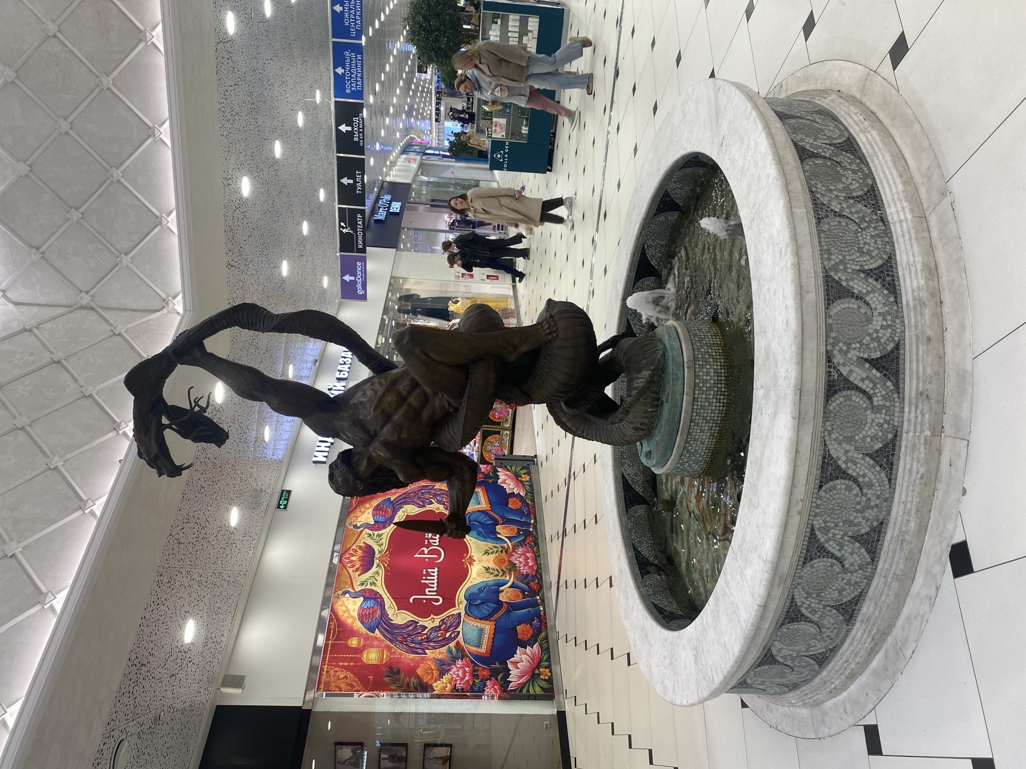 This indoor shot shows a portion of a shopping mall's interior in Yekaterinburg, Russia. The floor is white tile with black accents. A sculpture of a man entwined with a serpent stands near a circular fountain with cascading water. Behind the sculpture is an advertisement with the text "India Baz." A few pedestrians walk in the background, passing storefronts with visible signage. The ceiling has a patterned texture with recessed lighting. Directional signage is visible, indicating parking and restroom facilities.