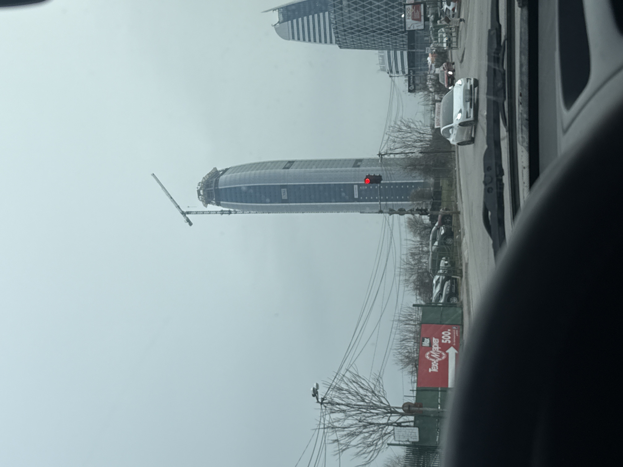 The image depicts a view from inside a vehicle, looking out at a cityscape in Sofia, Bulgaria. Dominating the scene is the Capital Fort skyscraper, still under construction with a crane visible near its top. Adjacent to Capital Fort, there is another, more completed skyscraper. A white car is visible in the foreground, on what appears to be a road alongside the building. A red sign for "Max Text Mapier 500" is also visible near some trees. The sky is overcast.