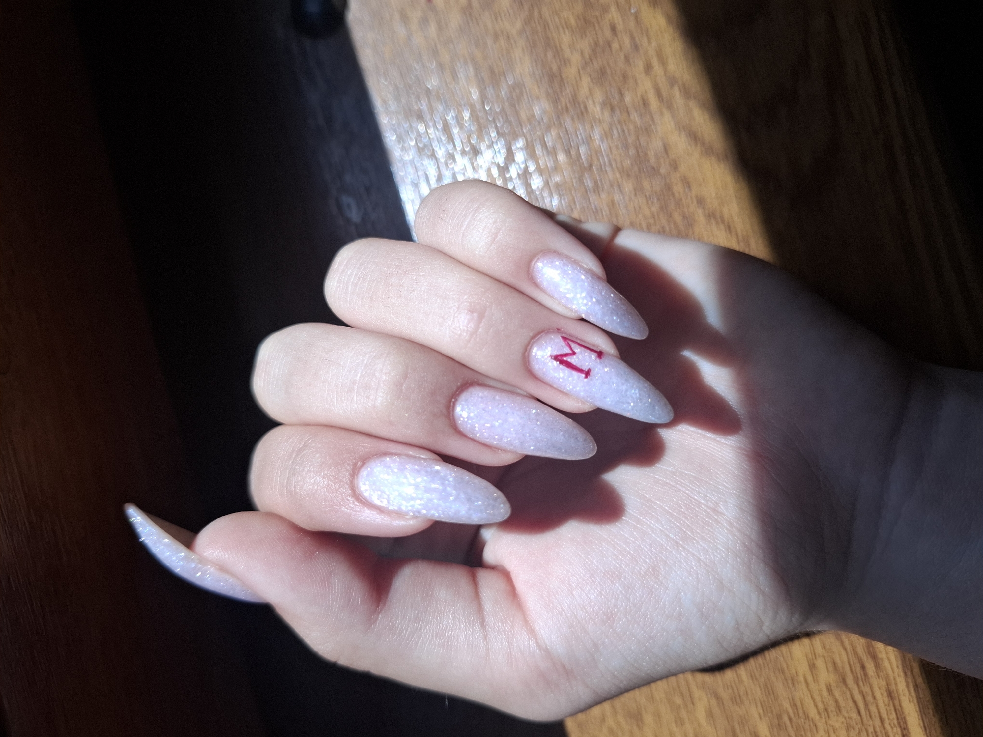 The image shows a close-up of a person's hand, featuring long, pointed nails with a pearlescent white polish and glitter. One of the nails, on the ring finger, has a small, red letter "M" painted on it. The hand is resting on a wooden surface, and there is a shadow cast across the scene, suggesting it is outdoors or near a window. The background is dark and out of focus. The image does not contain any elements that would indicate a location in the city of Zamfir, Bulgaria.