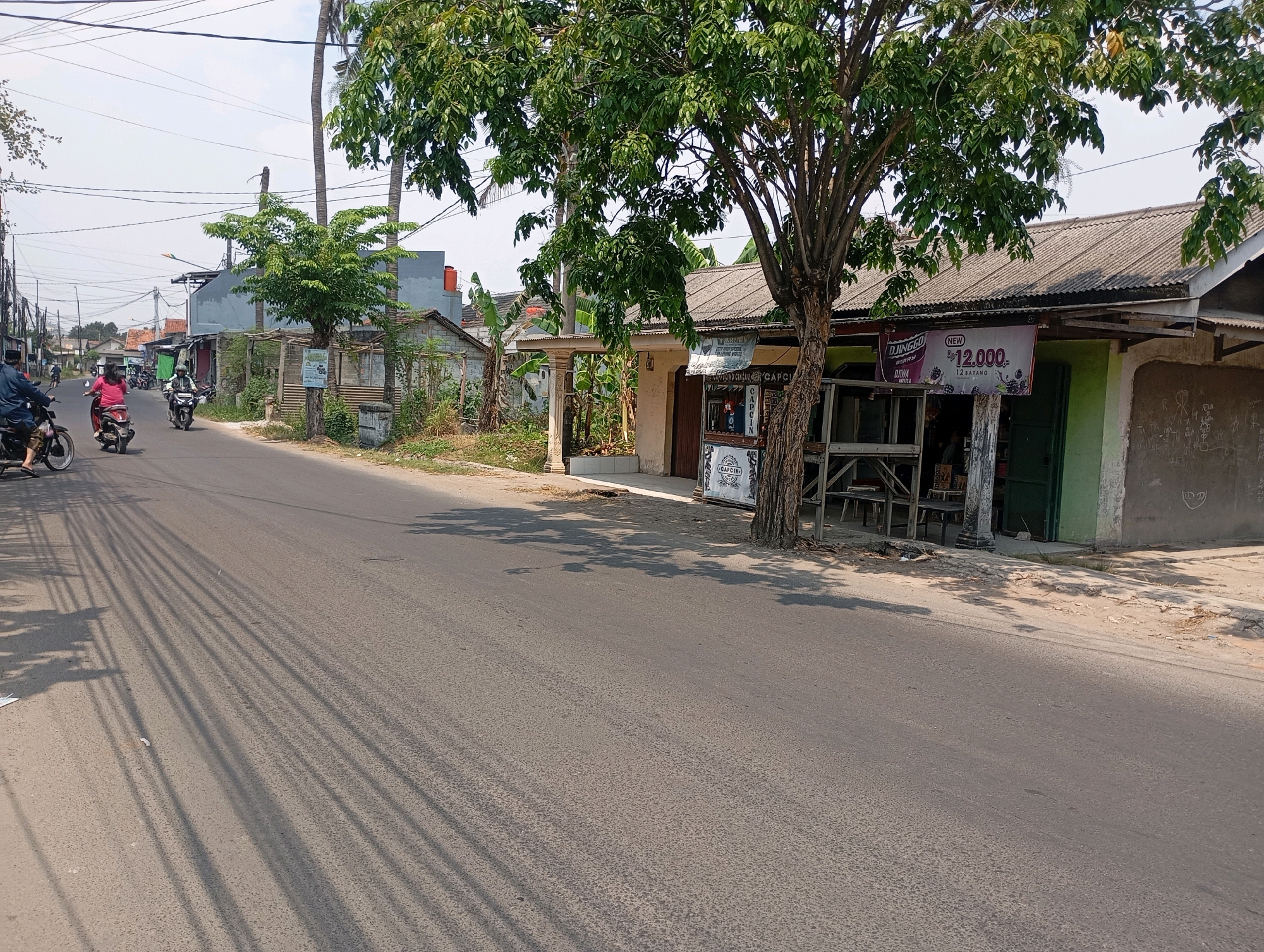 The image depicts a street scene in Babelan, Indonesia, with a road running diagonally from the bottom left to the top right. On the right side of the road, there are several buildings with corrugated metal roofs. One building has a green door and a sign that advertises "DJINEGGO BERRY" with "Rp 12.000." A large tree with dense green foliage stands in front of the buildings, casting shadows on the road. Several power lines are visible crisscrossing the sky.
On the left side of the road, a few people are riding motorcycles. One person in a blue shirt is on a motorcycle in the foreground, and another person in a pink shirt is on a motorcycle further up the road. Several other motorcycles and people are visible in the distance, suggesting ongoing traffic. The environment appears to be a residential or commercial area with a mix of houses and small businesses. The road surface is asphalt. The sky is bright and appears to be daytime, with the sun casting strong shadows.