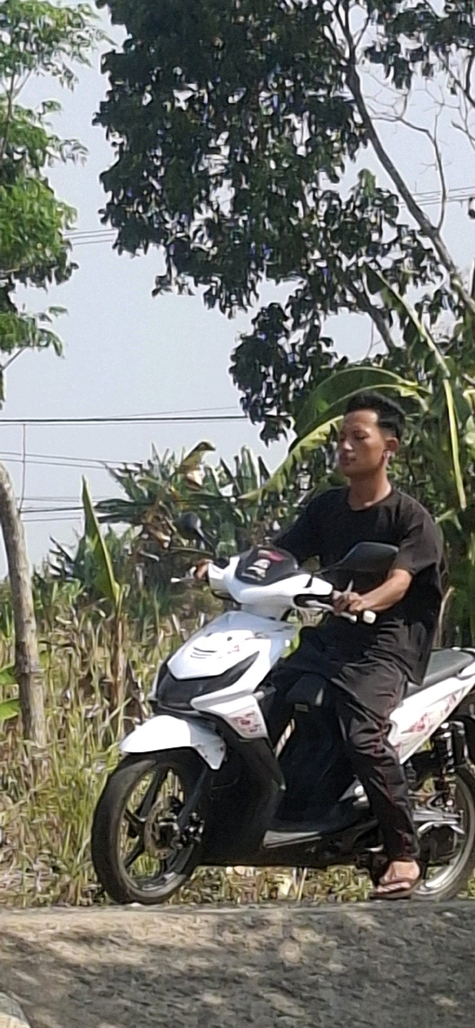 In Cibinong Hilir, Indonesia, a young man with dark hair and a black t-shirt sits on a white scooter. He wears black pants and sandals. The scooter has red and white decals. He is positioned on a rough, gray surface, possibly a road or path, with lush green vegetation and trees forming the backdrop under a pale sky. Power lines are visible in the distance.