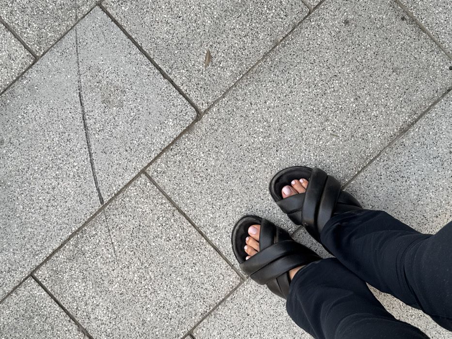 The image displays a low-angle perspective of a person's lower legs and feet, likely situated on a paved surface in Dubai, United Arab Emirates. The individual wears black, loose-fitting trousers and black, padded, cross-strap slide sandals. The toes of both feet are visible, each with a light pink nail polish application. The ground consists of light grey, speckled concrete or terrazzo tiles, laid in a pattern of rectangular and trapezoidal shapes with visible grout lines. A thin, dark linear mark extends across several tiles in the upper central area of the frame, and a small, dry, brown leaf is present on the pavement in the lower right quadrant. No other individuals or significant environmental features are visible.