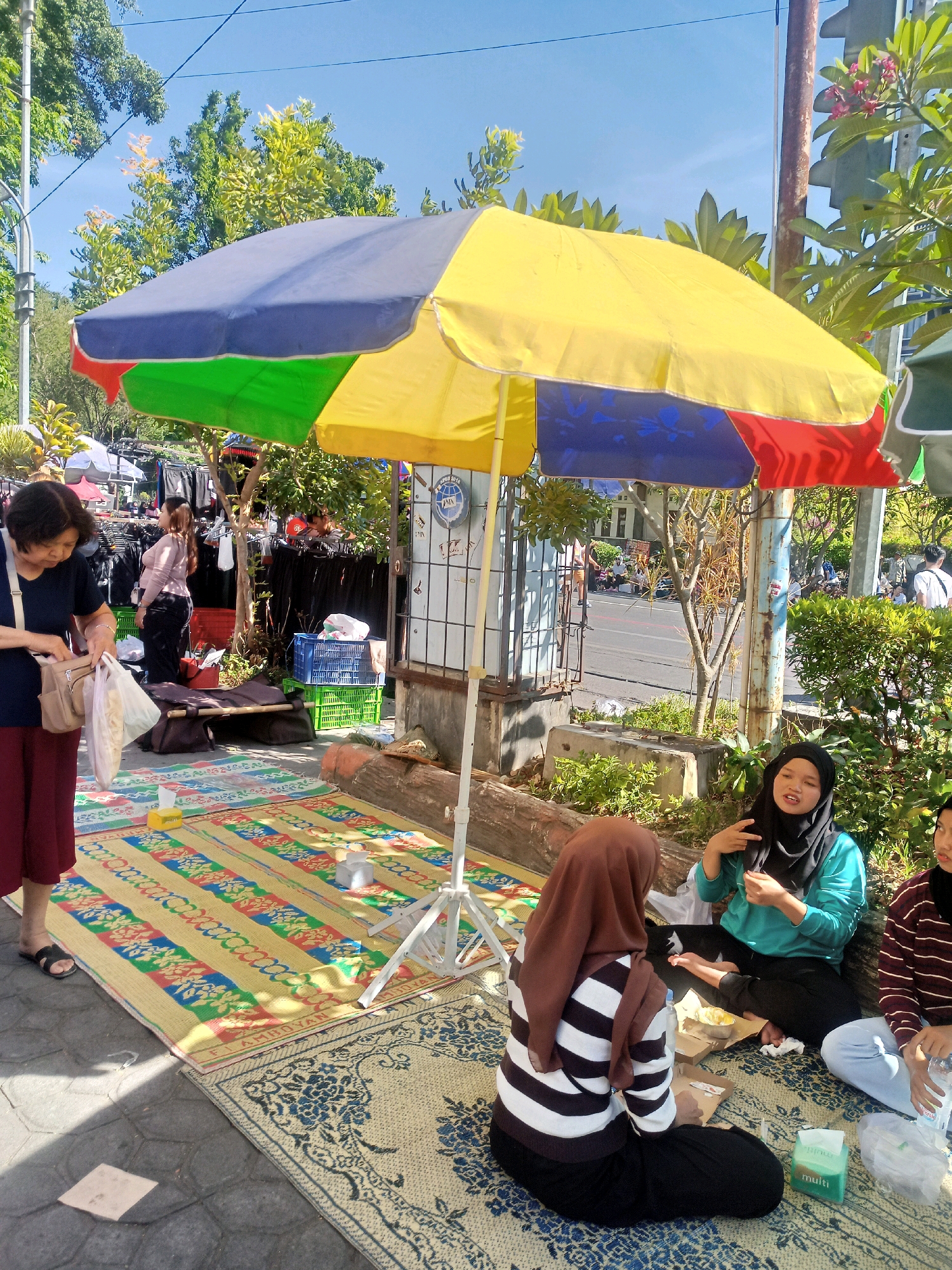 In the city of Surakarta, Indonesia, a sunny outdoor scene unfolds with people gathered under a large, colorful umbrella. The umbrella, divided into sections of yellow, red, green, and blue, provides shade over a street market. Several individuals are seated on woven mats laid out on a paved area adjacent to a road.
In the foreground, a woman with short dark hair, wearing a dark blue top and a maroon skirt, is seen from the waist down, holding a plastic bag and a purse. To her right, three young women are seated on the mats. One, wearing a teal top and a black hijab, gestures with her hands while speaking. Another, in a brown hijab and a black and white striped sweater, is seen from the back. The third woman, on the far right, is partially visible and appears to be wearing a striped long-sleeved shirt and light-colored pants. Food containers and a box of tissues are scattered on the mats.
Behind the seated individuals, a market stall displays clothing on racks. Various items, including bags and baskets, are visible in the background. A metal fence and a tree stand near the stall, with a street and more trees in the distance. The overall environment suggests a vibrant street market on a clear day.