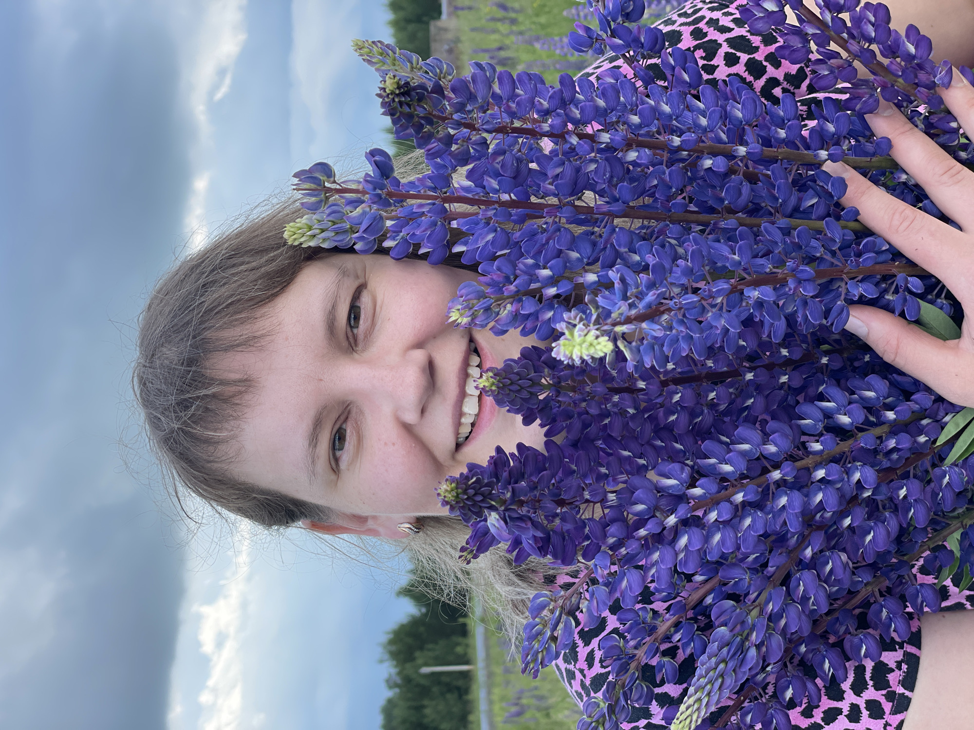 In Прикамский, Russia, a woman with light brown hair and bangs smiles as she holds a large bouquet of purple lupine flowers. She is wearing a pink and black leopard-print top. The background shows a field of lupine flowers and a cloudy sky.