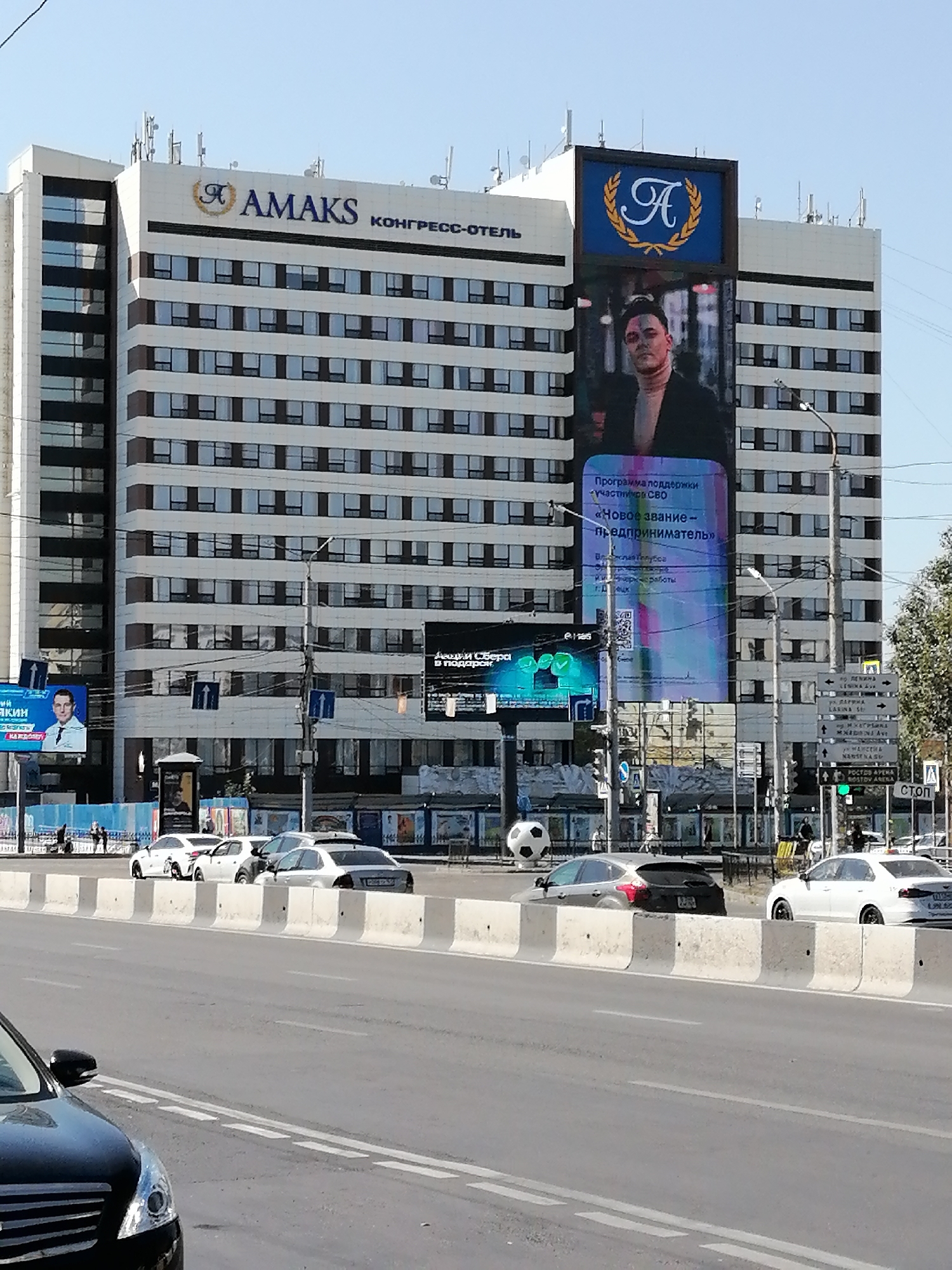 The image shows a wide, multi-lane road in Rostov-on-Don, Russia, with a large, modern hotel building in the background. The hotel, identified by a sign reading "AMAKS КОНГРЕСС-ОТЕЛЬ", is a tall, white building with many windows and a prominent digital billboard. The billboard displays an advertisement featuring a man in formal attire and text in Russian. On the road, several cars are visible, including a black sedan in the foreground and white cars in the middle distance. A concrete barrier separates the road from a sidewalk area where people are walking and a large soccer ball sculpture is present. Traffic lights and street signs are also visible, indicating a busy urban environment. The sky is clear and blue, suggesting a sunny day.