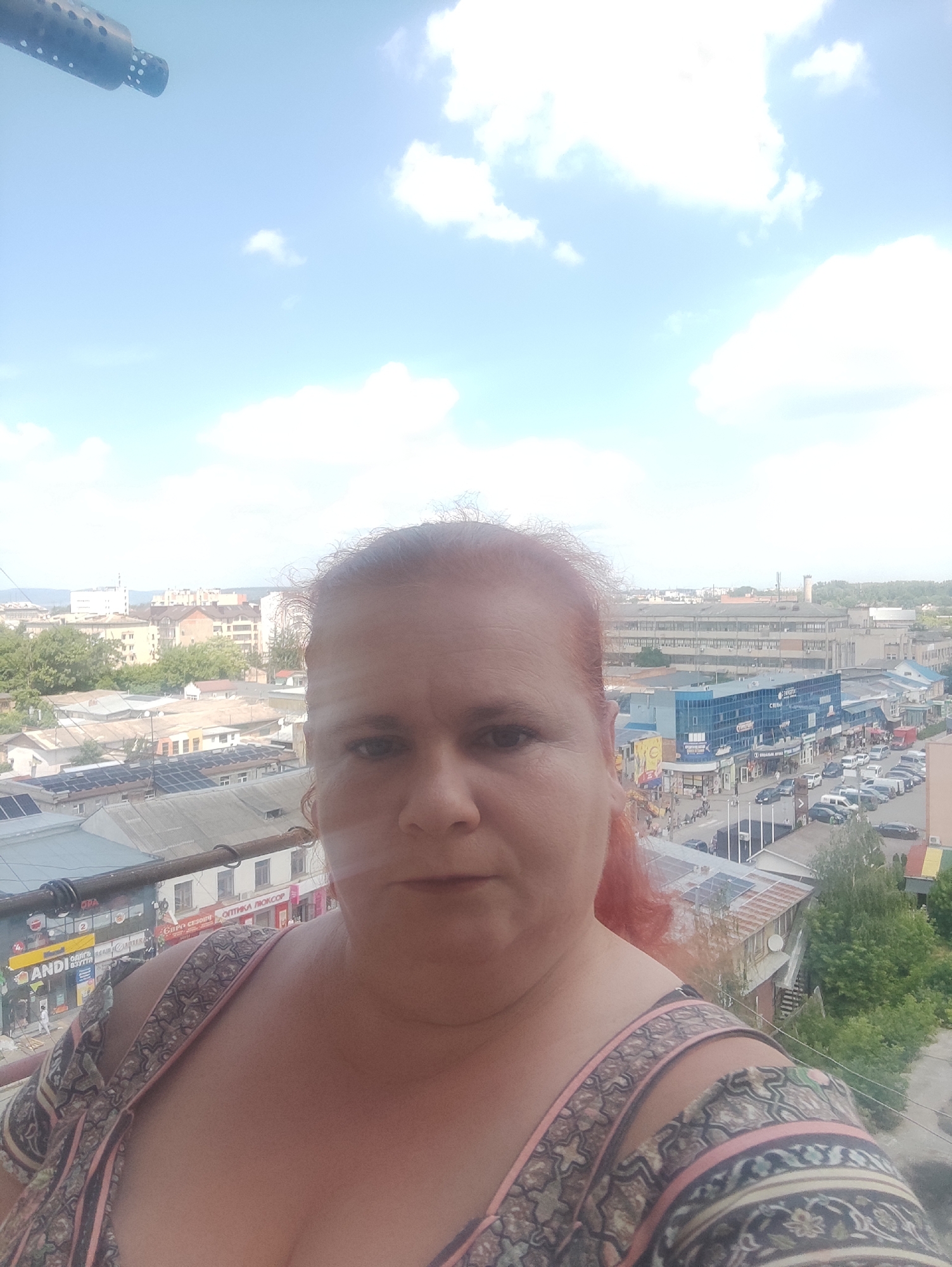 A selfie captures a woman with red hair and a light-skinned complexion from the chest up. She is wearing a low-cut top with a pink, beige, and black patterned design. Her shoulders and cleavage are visible. The background depicts an aerial view of a city. Buildings of various sizes and architectural styles are visible, including some with flat roofs and others with tiled roofs. Several streets are lined with cars. Some buildings have visible signage, such as "ANDI," "ОПТИКА ЛЮКСОР," and "БИРО СЕЗОНИ." Green trees are interspersed throughout the urban landscape. The sky above is blue with scattered white clouds. In the upper left corner of the image, a portion of a metallic object, possibly a railing or part of a structure, is visible. The scene is set in the city of Chernivtsi, Ukraine.