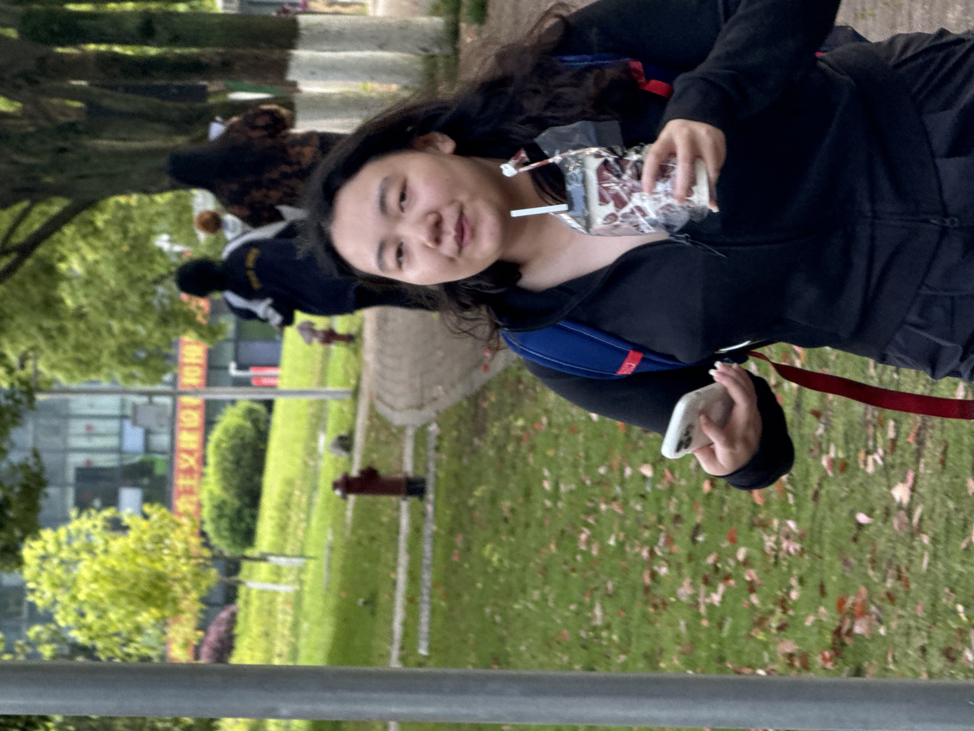 A young Asian woman is walking outdoors, holding a beverage with a straw in her right hand and a white smartphone in her left. She is wearing a black zip-up hooded jacket, and a blue backpack with red straps is visible on her shoulders. She is facing the camera with a slight smile.
The background consists of a grassy area covered with fallen leaves, along with a sidewalk and a building in the distance. A red fire hydrant is visible on the grass. Text in Chinese characters can be seen on a sign on the distant building. Based on the characters and their context in the image, the location can be identified as Jiangjin, China. Other people can be seen in the background.