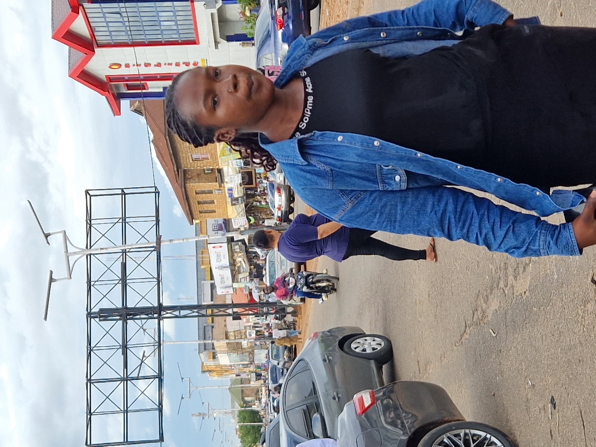 The image depicts a street scene in Jos, Nigeria, under an overcast sky. In the foreground, a young Black woman with braided hair is the primary subject, facing the camera. She is wearing a blue denim jacket over a black t-shirt with white lettering that reads "Soipme Acne S". Her expression is neutral, and she has a nose ring. Behind her, traffic is visible on a paved road. Several cars, including a grey sedan and a dark blue sedan, are parked or moving. A man rides a black motorcycle in the middle ground, wearing a pink shirt. Other pedestrians and more vehicles are present further down the street, along with buildings lining the road. One prominent building on the right features red-trimmed white architecture with blue window frames. A large metal billboard structure stands in the background. The overall environment suggests a bustling urban area.