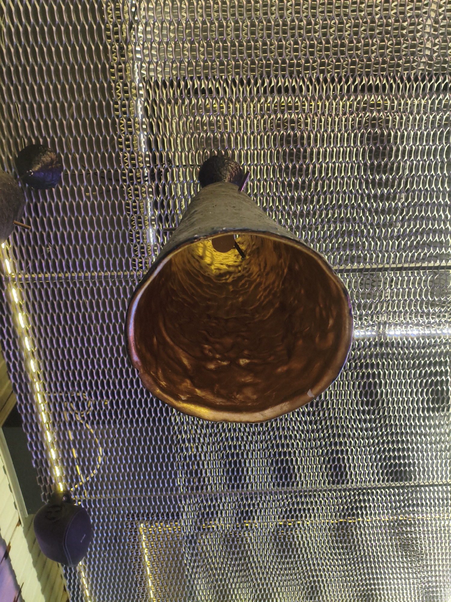 An overhead shot captures a conical pendant light fixture with a hammered copper interior, illuminated by a visible light bulb within. The fixture hangs from a gray, diamond-patterned metal mesh ceiling, which is subtly lit by strings of yellow LED lights. Several dark, spherical objects, likely speakers, are also suspended from the ceiling by thin wires, one prominently visible in the lower left quadrant. The background appears to be a textured wall or structure with muted lighting. The scene is devoid of any people and provides no specific indicators of its location within Kremenchuk, Ukraine.