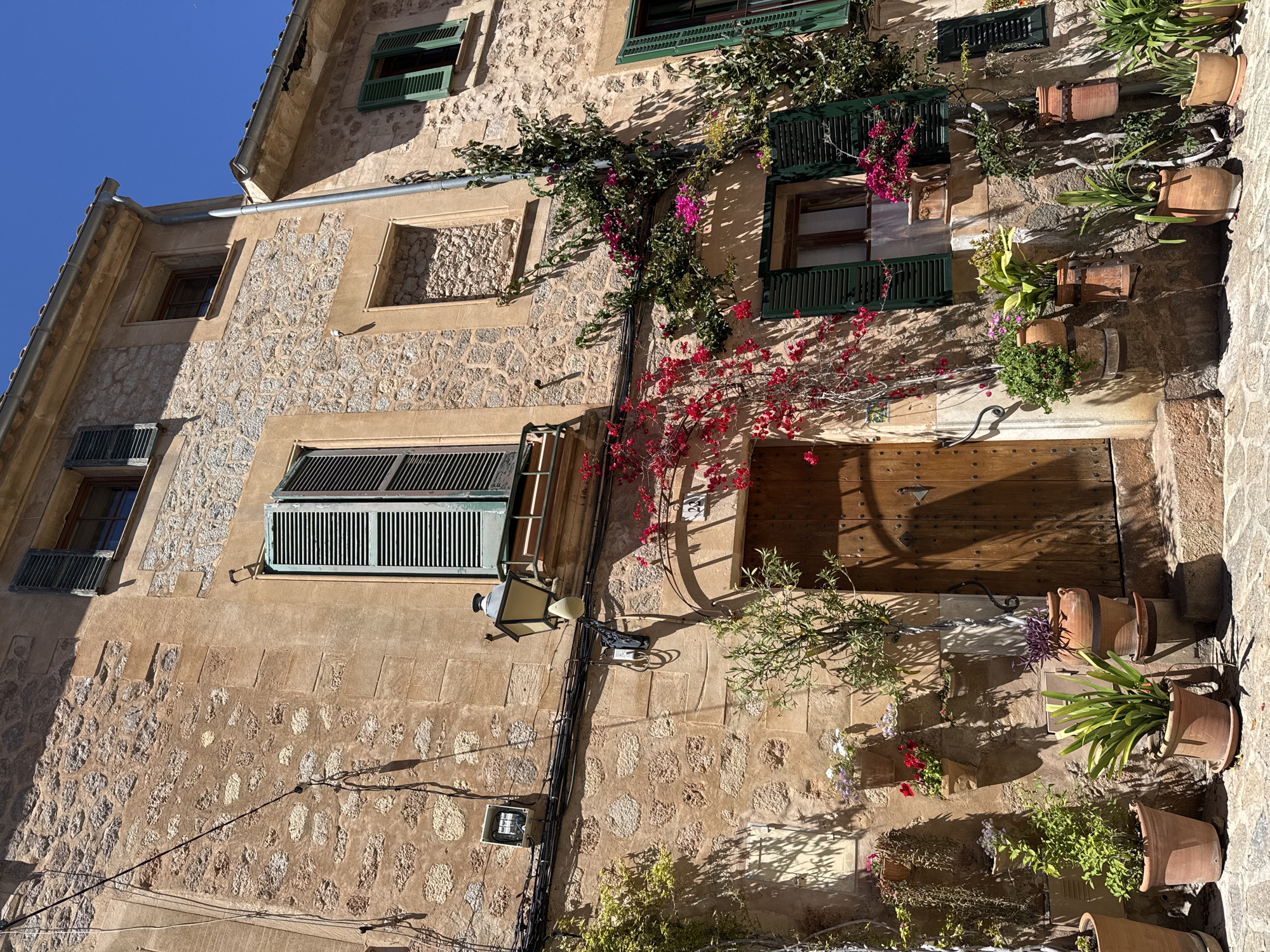 The image shows a building in Valldemossa, Spain, characterized by stone walls and a wooden door. Green shutters adorn the windows, some of which are partially open. Bougainvillea climbs the facade. A street lamp is affixed to the wall near the door. Clay pots with plants decorate the exterior. Electrical wires are visible along the wall. The image is taken during daylight.