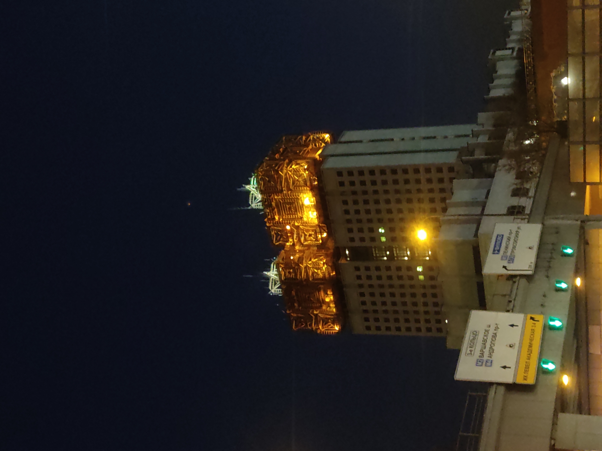 The image depicts a nighttime scene in Moscow, Russia, featuring a large building with a distinctive golden, ornate structure at its top. The rest of the building appears to be a standard high-rise with a grid of lit windows. The sky is a dark blue. The street level includes road signs. One sign reads "3-e Koltso" and points toward the Warsaw Highway and Andropova Avenue. Another sign refers to the Level Akademicheskaya 34 residential complex. The road signs are backlit with green lights. There are lights on the surrounding structures as well.
