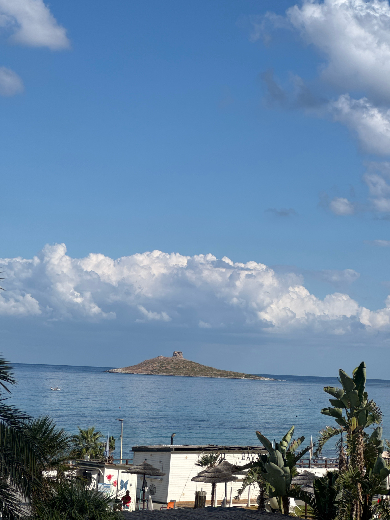 The image depicts a coastal scene with a small island in the distance, featuring a stone structure atop it. The foreground is occupied by a white building with a "BAR" sign, palm trees, and thatched umbrellas, suggesting a beachside establishment. In the middle ground, the clear blue sea is visible, with a small white boat navigating the water. The sky is a bright blue, with scattered white clouds, indicating a clear day. The location is identified as Capaci, Italy.