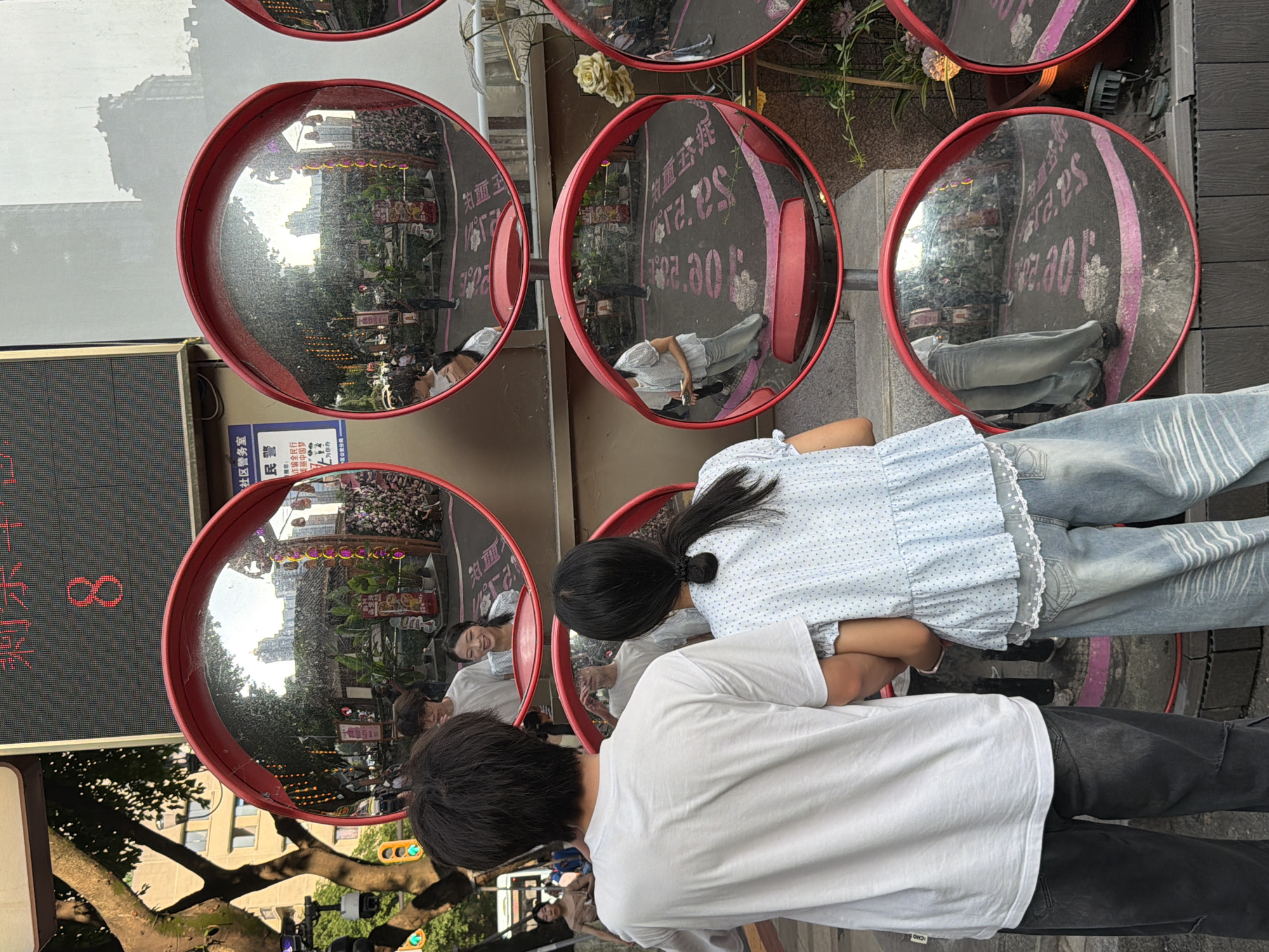 In Yuzhong District, China, two individuals, a male and a female, stand facing away from the camera, their backs to the viewer. The male, on the left, wears a white t-shirt and dark pants. The female, on the right, is dressed in a light blue, ruffled shirt and light blue jeans. They appear to be a couple, standing close together, possibly on a street or public walkway. The scene is dominated by a series of convex mirrors, mounted on poles, reflecting the surrounding environment. These mirrors show a street scene with other people, buildings, trees, and what appears to be a digital display board in the background showing the number "8" in red. The ground beneath the mirrors has pink markings, possibly indicating a crosswalk or designated area. The overall atmosphere suggests an urban setting during daytime, with a mix of natural and man-made elements.