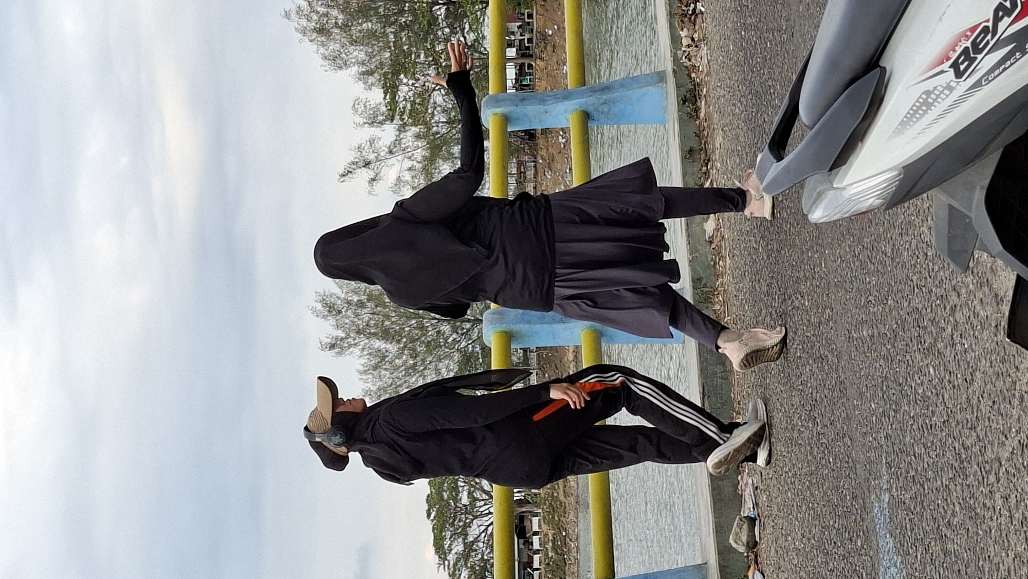Two individuals, likely female, are captured from behind, walking on a paved walkway beside a body of water. The individual on the left wears a black hijab, a beige baseball cap, headphones, a black hooded jacket, and black track pants with white stripes. They are holding an orange object. The individual on the right wears a black hijab, a black long-sleeved shirt, and a dark grey pleated skirt over black leggings. Both are wearing athletic shoes. The walkway is bordered by a yellow railing with blue posts, separating it from the water. Trees line the far bank of the water, with some structures visible in the background. The ground on the walkway is paved with coarse gravel. In the foreground, the rear section of a white and black motorcycle is visible. The sky is overcast. This scene is consistent with a public recreational area or embankment, possibly in Lhokseumawe, Indonesia, given the cultural attire and common elements of urban landscaping in such regions.