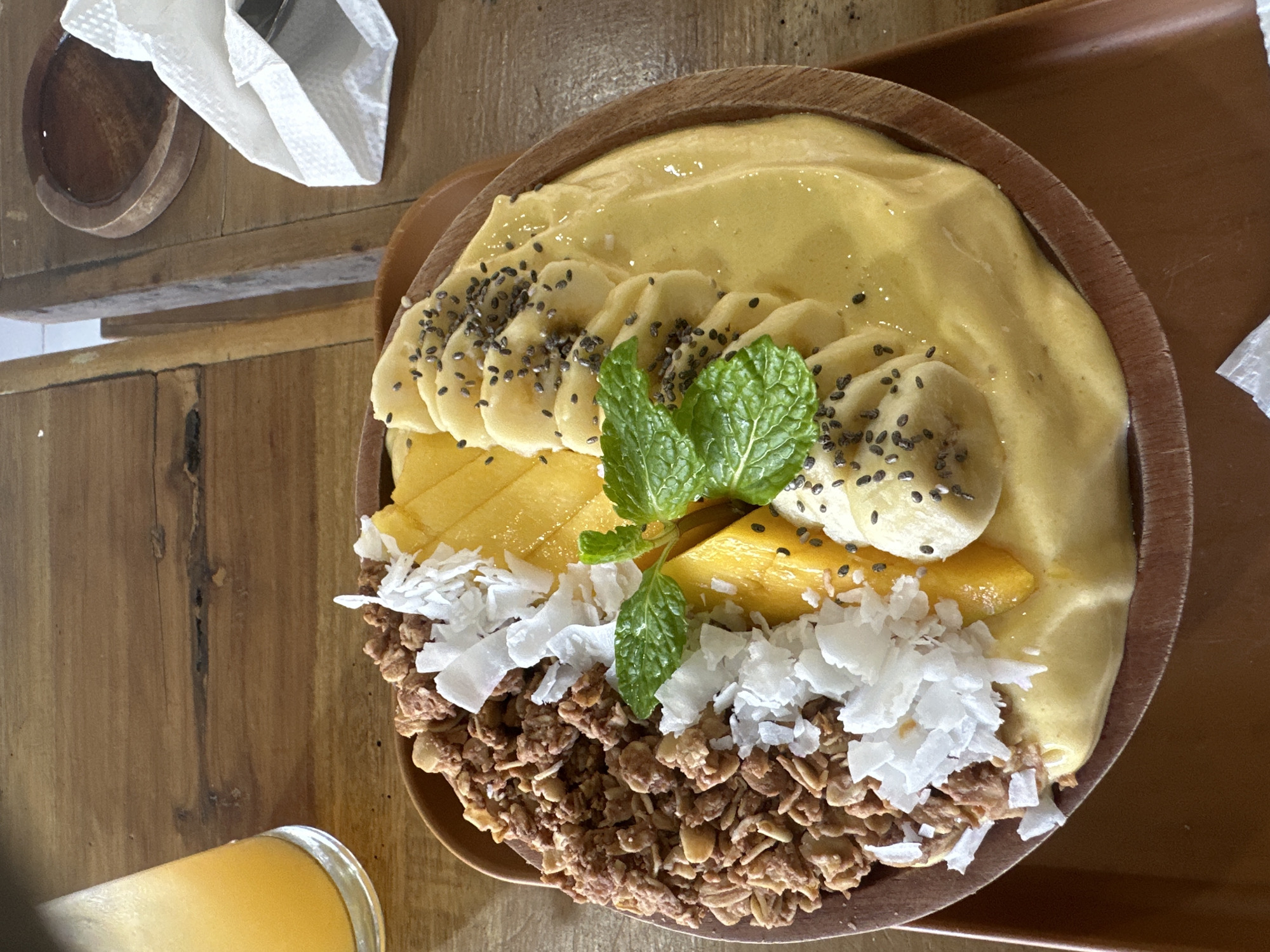 An overhead shot captures a vibrantly presented smoothie bowl on a wooden table, likely in Kuta Selatan, Indonesia. The bowl, made of dark wood, is filled with a creamy yellow smoothie topped with a generous serving of granola, shredded coconut, sliced mango, and thinly sliced bananas. Chia seeds are sprinkled over the bananas, and fresh mint leaves add a touch of green to the arrangement. To the left of the bowl, a glass of orange juice is partially visible. The wooden table shows a natural grain pattern, and a white napkin is folded to the right of the bowl.