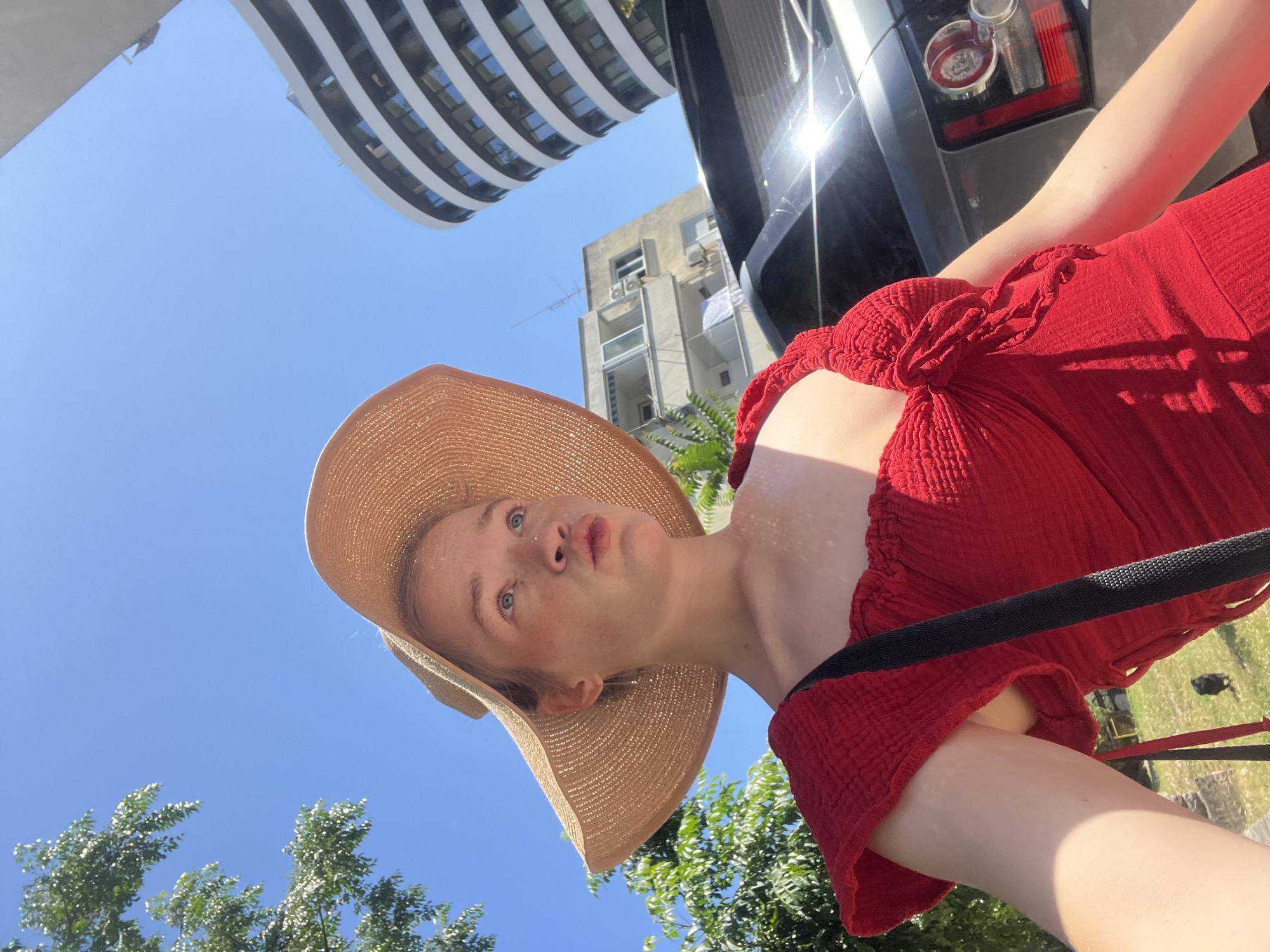 A low-angle, sunlit selfie in Budva, Montenegro, features a fair-skinned individual, likely female, wearing a wide-brimmed straw hat and a textured red off-the-shoulder top. Her face, adorned with freckles, is turned slightly upwards and to her left, with her lips subtly puckered. A thin silver earring hangs from her left ear, and a black strap crosses her right shoulder. The background, under a clear blue sky, includes green tree foliage, a patch of grass with a small dark animal, a modern curved white building, an older apartment building with balconies and visible laundry, and a parked dark gray SUV reflecting intense sunlight.