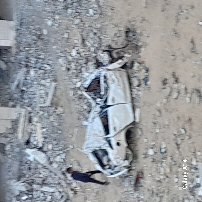 The image depicts a scene of extensive structural damage in Khan Yunis, Palestinian Territory. The foreground and midground are covered in a dense layer of rubble, consisting of broken concrete, rebar, and debris. Centrally located is a heavily damaged white four-door sedan, its roof completely crushed, body panels severely dented and distorted, and front and rear sections mangled. The vehicle appears partially submerged in a sandy-colored dirt mixed with concrete fragments. In the background, indistinct, partially standing structures made of concrete are visible amidst further rubble. On the left side of the frame, a single individual, dressed in dark clothing, walks towards the right across the debris-strewn ground. There are no direct interactions between the person and the vehicle or other subjects.