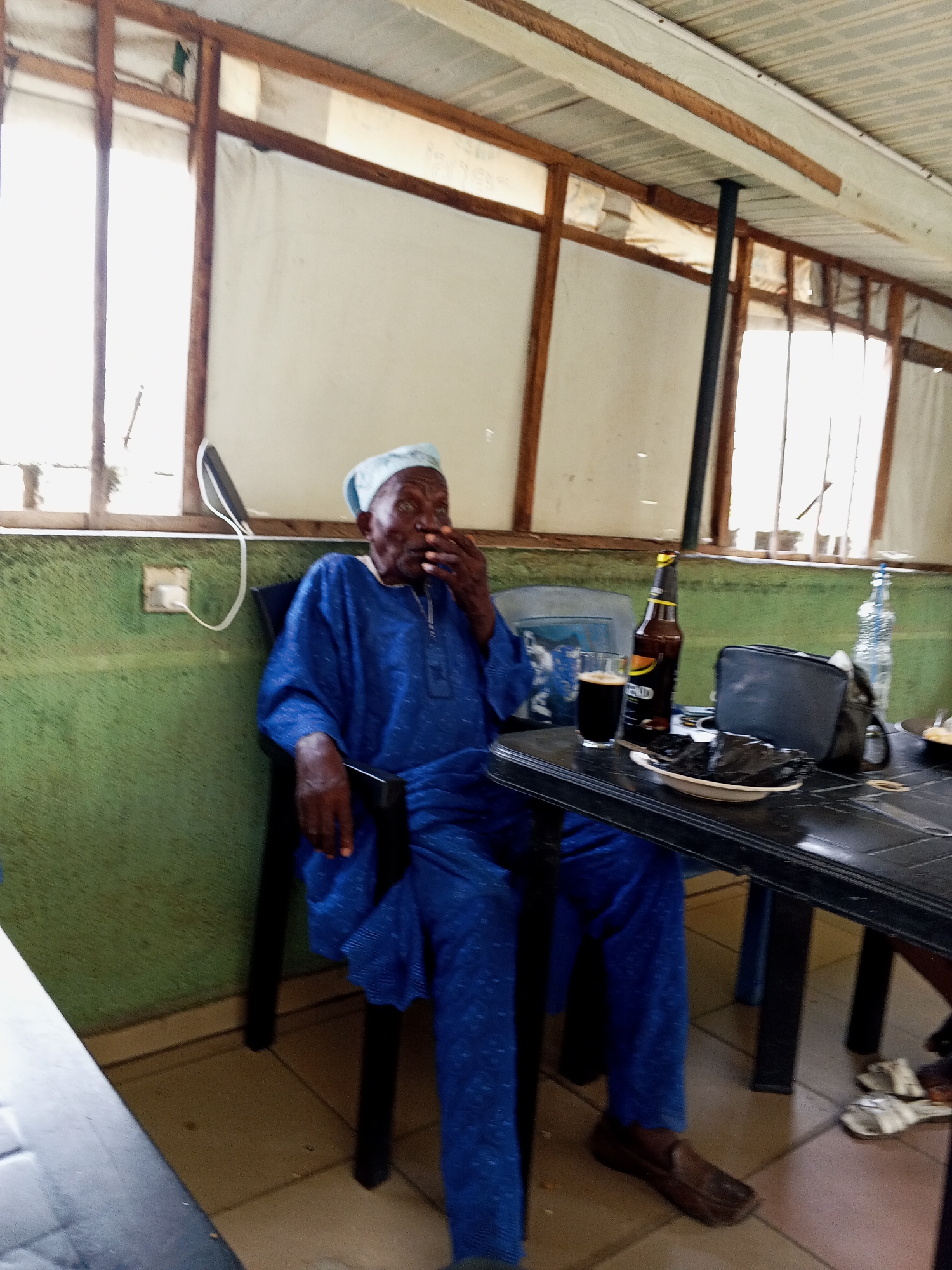 An older man, dressed in a blue traditional garment (agbada-style) and a light blue cap, is seated on a black chair at a black plastic table. His right hand is raised to his mouth or chin, and he appears to be looking to the right. On the table in front of him are a glass of dark liquid (likely stout), a brown beer bottle with the brand "LEGEND" visible, a small black pouch, and a white plate holding dark, wrapped food items, possibly street food. A clear plastic water bottle is also partially visible on the table.
The setting appears to be an indoor space, possibly a casual eatery or bar in Zango-ndaji, Nigeria. The walls are painted green in the lower section and lighter above. Windows with wooden frames are visible in the background, allowing bright daylight to stream through, indicating it is daytime. The windows are covered with some opaque material, possibly thin fabric or plastic sheeting. Wooden beams support the ceiling, which appears to be made of corrugated panels. A black metal pillar extends from the floor to the ceiling on the right side of the frame. A white phone charger cable is plugged into a wall socket on the green wall behind the man. The floor is tiled with light-colored tiles. White flip-flops are on the floor near the table to the right. The man wears dark brown slip-on shoes. He seems relaxed, perhaps enjoying a meal and a drink.