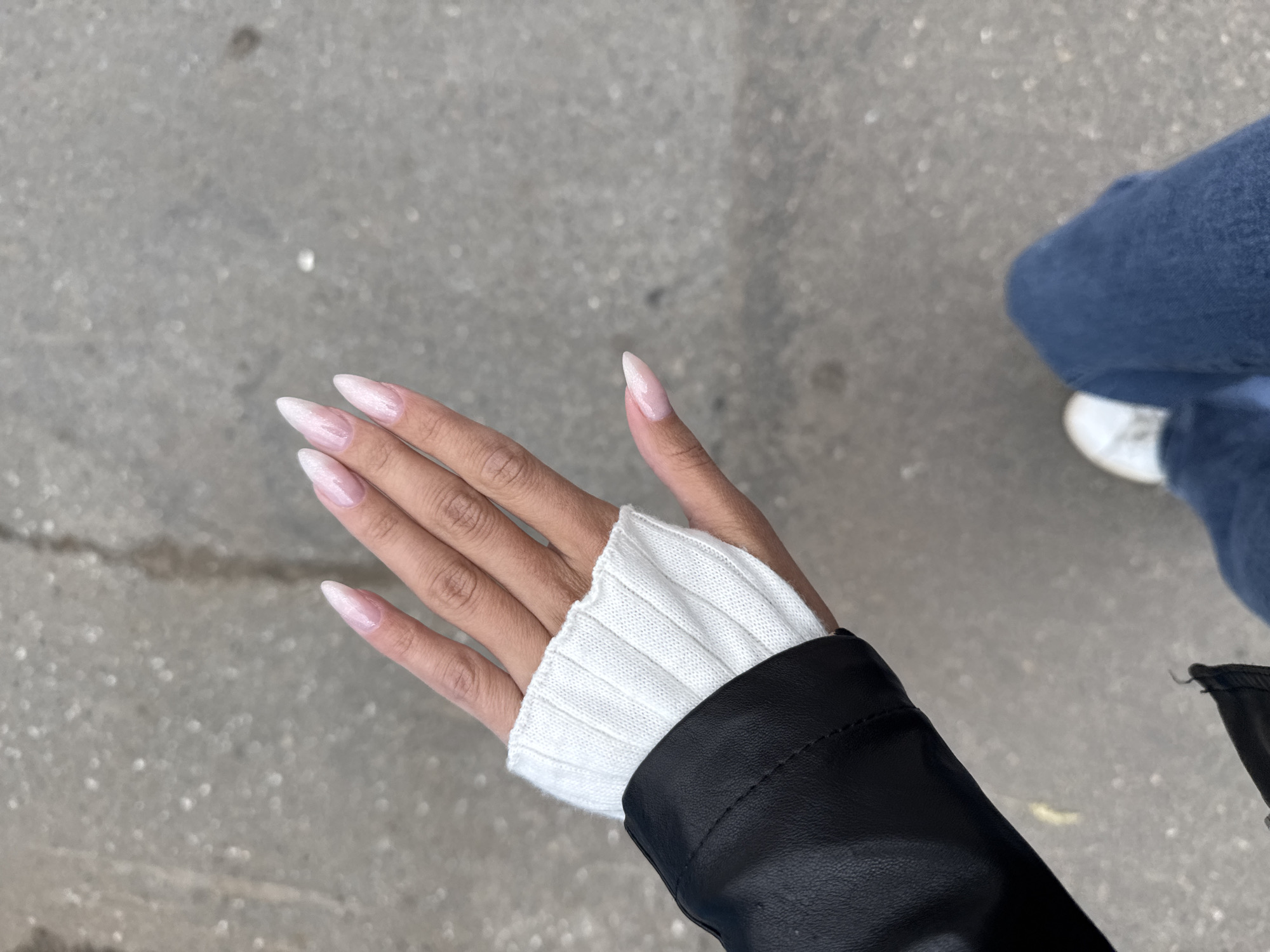The image shows a close-up view of a person's hand and arm from the shoulder down. The hand has long, almond-shaped nails with a pale pink, iridescent polish. The person is wearing a white ribbed sweater sleeve and a black jacket. The background is a gray, textured surface, likely asphalt, with some small white flecks. The lower part of the image shows a person's legs wearing blue jeans and white sneakers, suggesting they are walking on a street or sidewalk. There is no visible indication of the specific location being Izhevsk, Russia.