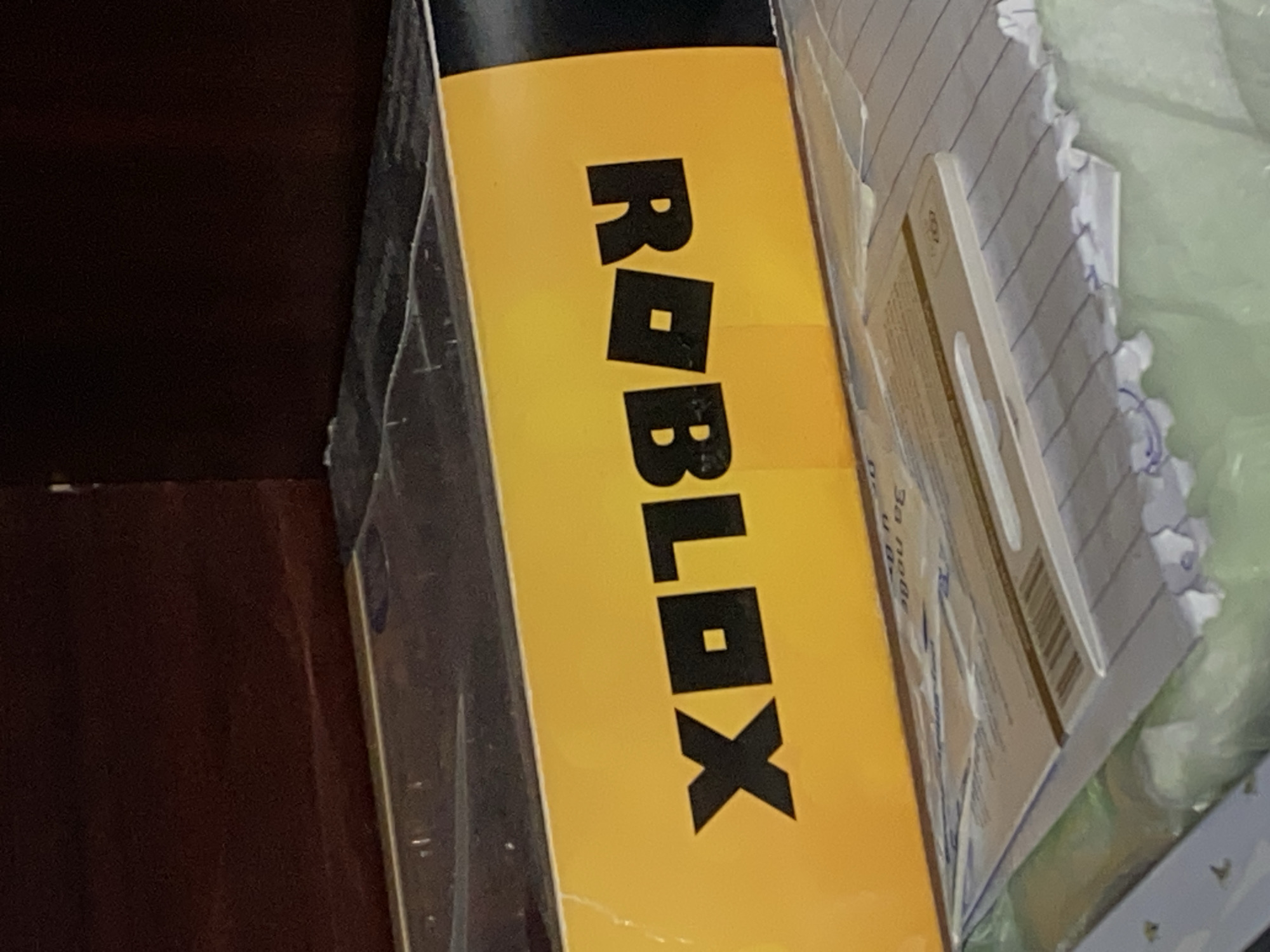 The image shows a yellow box with the word "ROBLOX" printed vertically in black, block letters. The box is placed adjacent to a stack of papers and possibly other objects. The background on the left shows a dark, solid-colored surface, possibly made of wood. The location is not discernible from the given image.