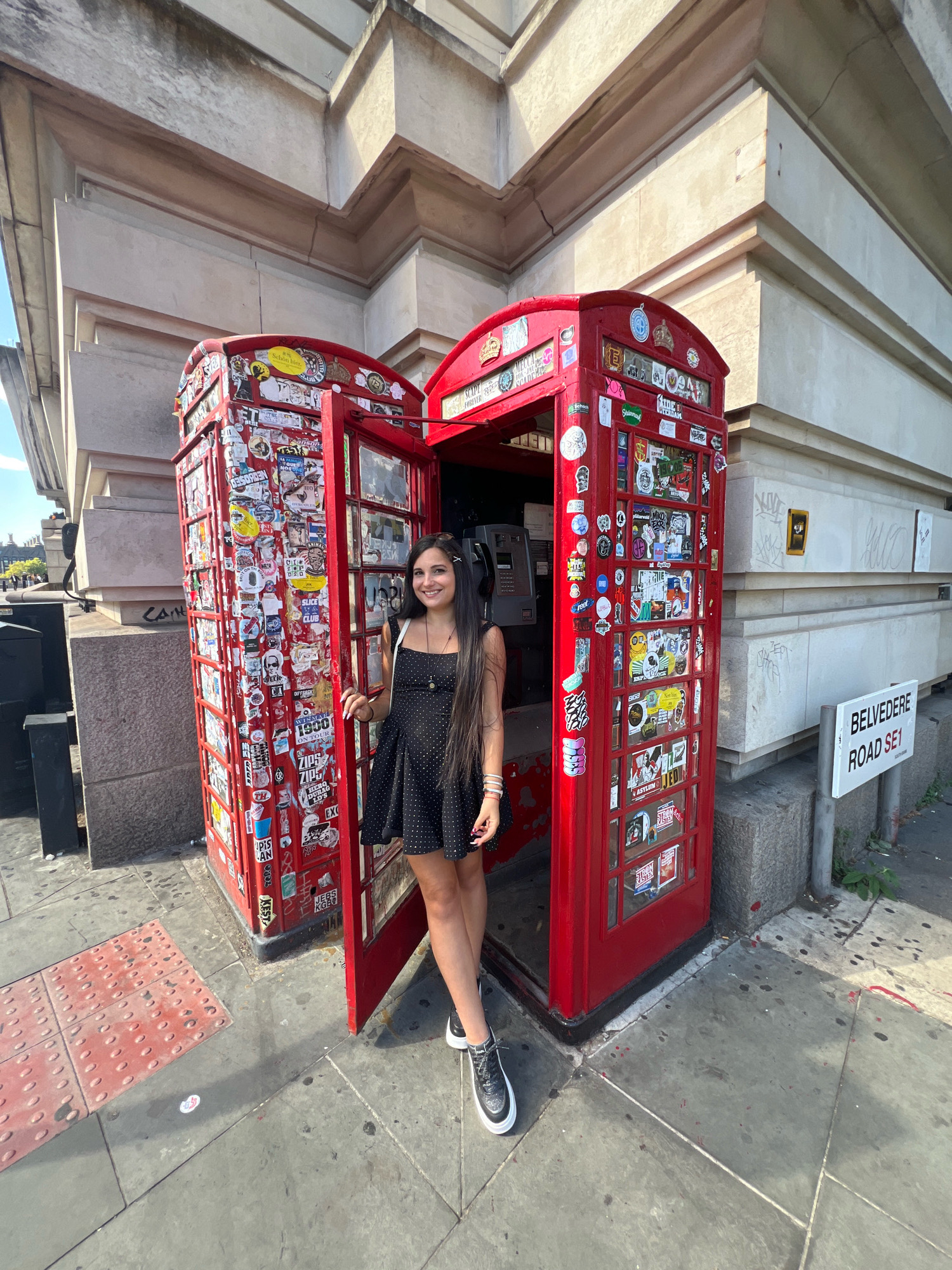 A young woman with long dark hair stands in front of a red, sticker-covered British telephone booth. She is wearing a black polka dot dress and black and white sneakers. The telephone booth is open, and she is standing with her legs crossed. Behind the booth is a large stone building with elaborate architectural details. A white street sign reads "BELVEDERE ROAD SE1" and is attached to a metal pole. The ground is paved with concrete and textured red tiles. The scene appears to be on a street in London, United Kingdom, likely near the South Bank of the River Thames given the Belvedere Road sign. The lighting suggests it is daytime, possibly late afternoon due to the long shadows.